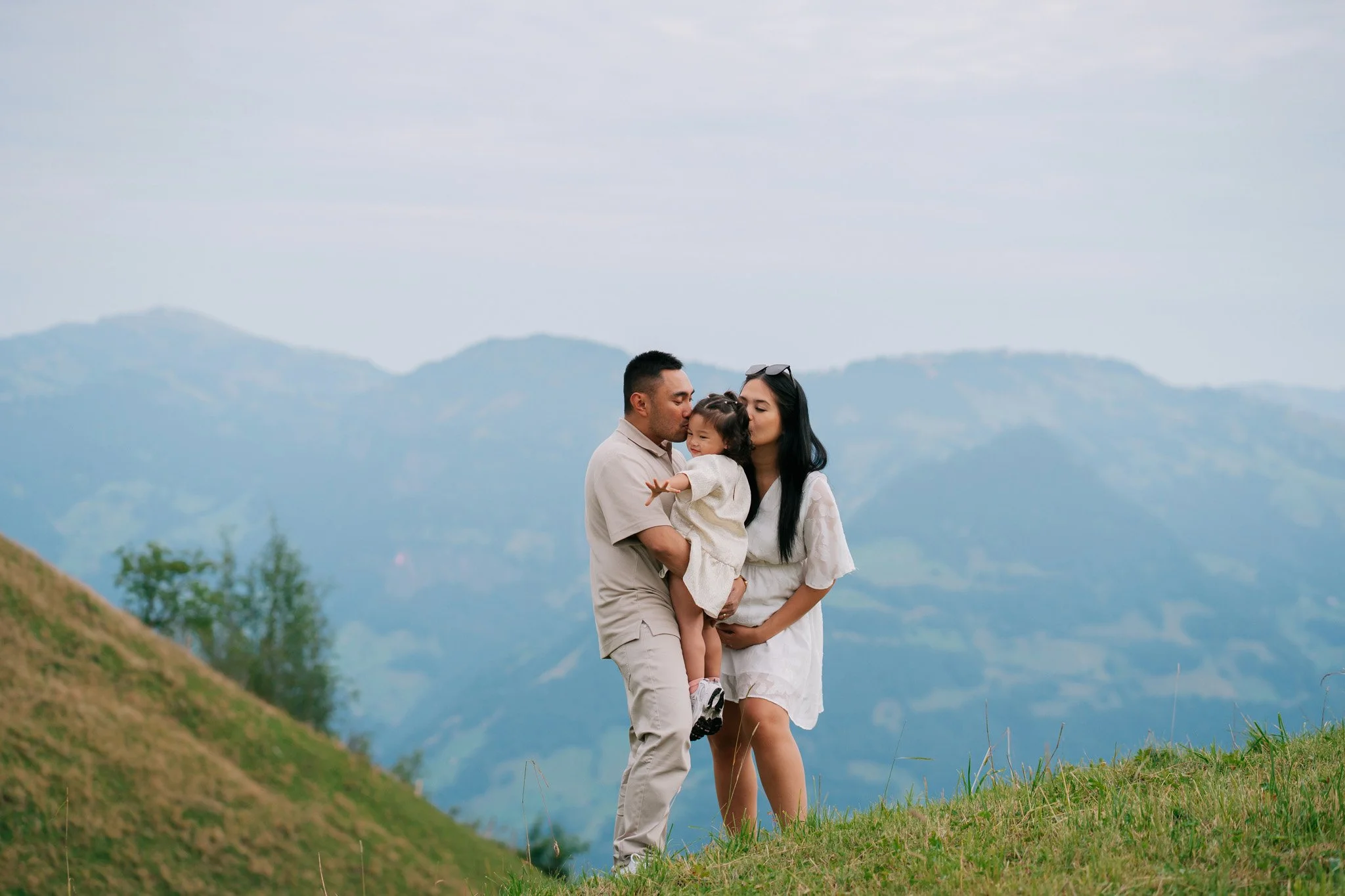 A family of three on a grassy hillside with mountains of Lucerne in the background, the father holding a young girl, and both parents kissing her cheeks. Family photoshoot in Switzerland.