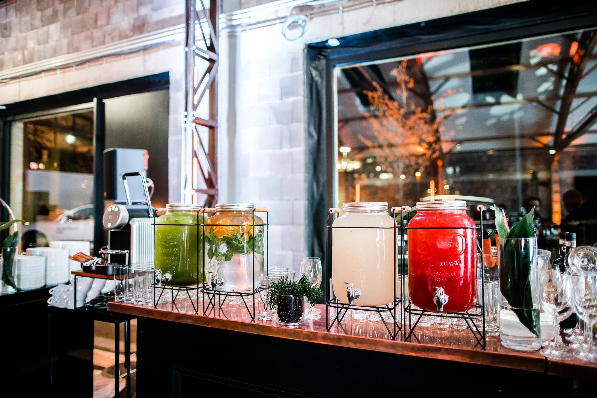 Four large glass beverage dispensers with spigots holding colorful drinks, arranged on a table at a night event. The drinks are green, orange, white, and red. There are glasses and a small potted plant nearby. Catering photographer Switzerland.