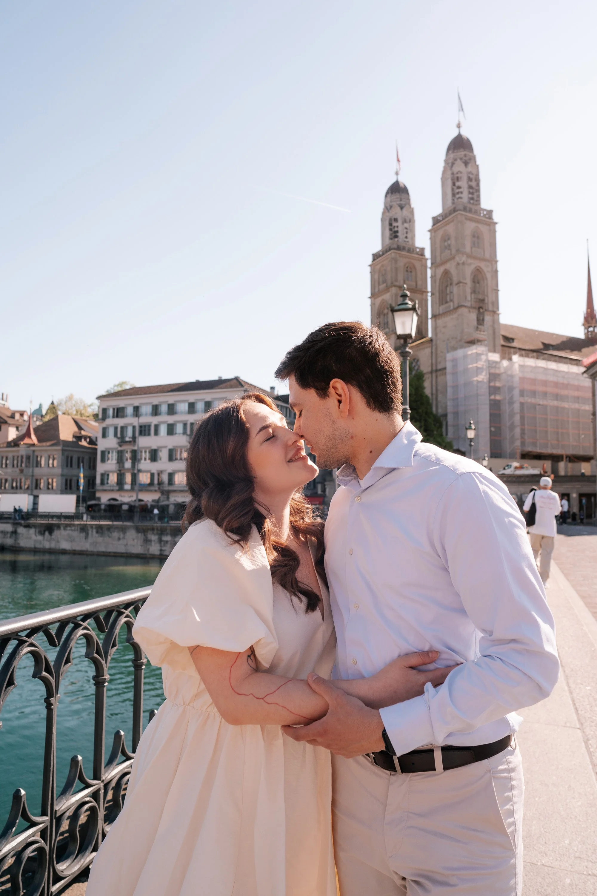 A couple embracing and about to kiss on a bridge in Zurich near a river with historic buildings and a church Grossmunster in the background.