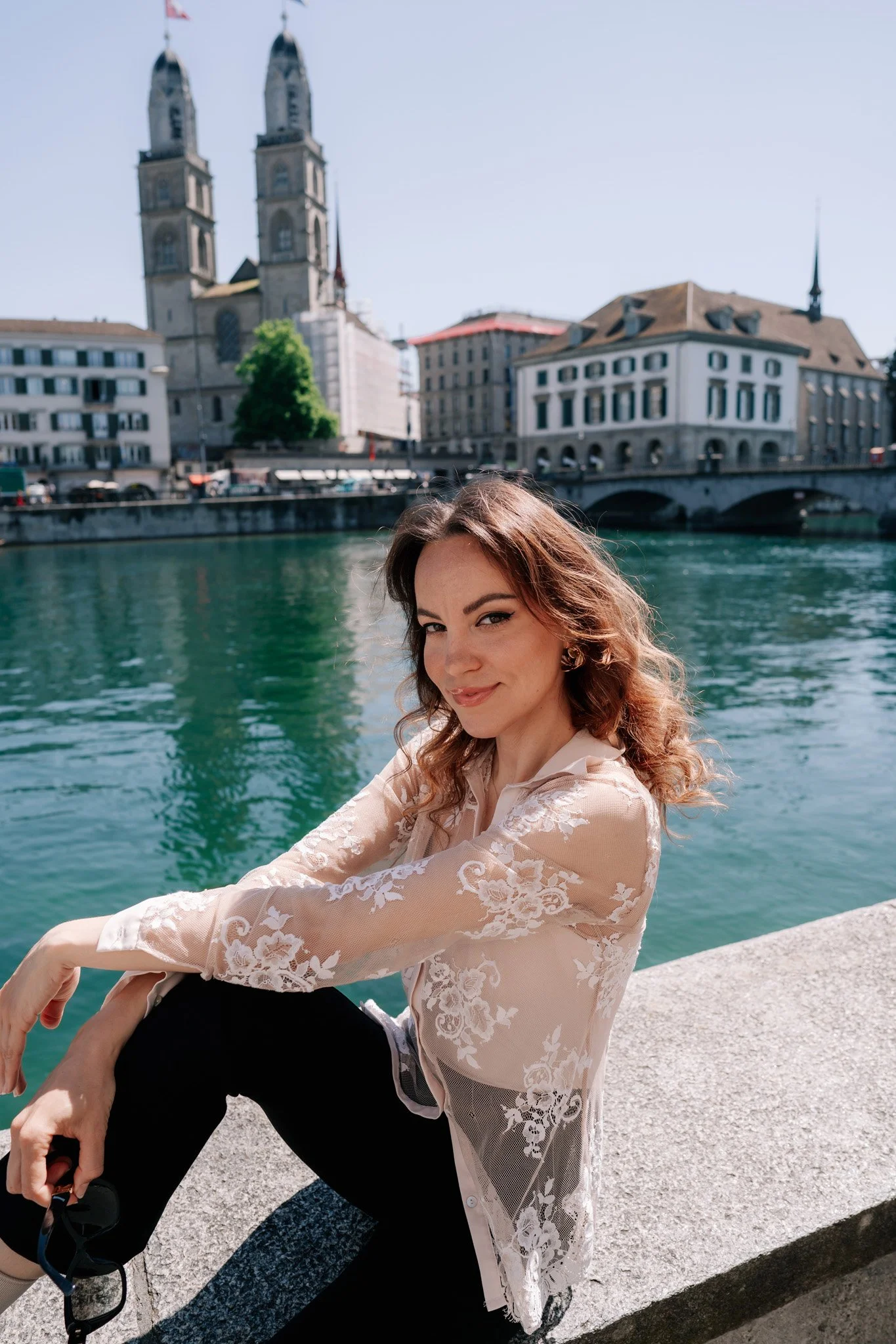 A woman sitting on a stone ledge near a river, with historic buildings of Zurich Switzerland and twin church towers in the background, smiling at the camera.