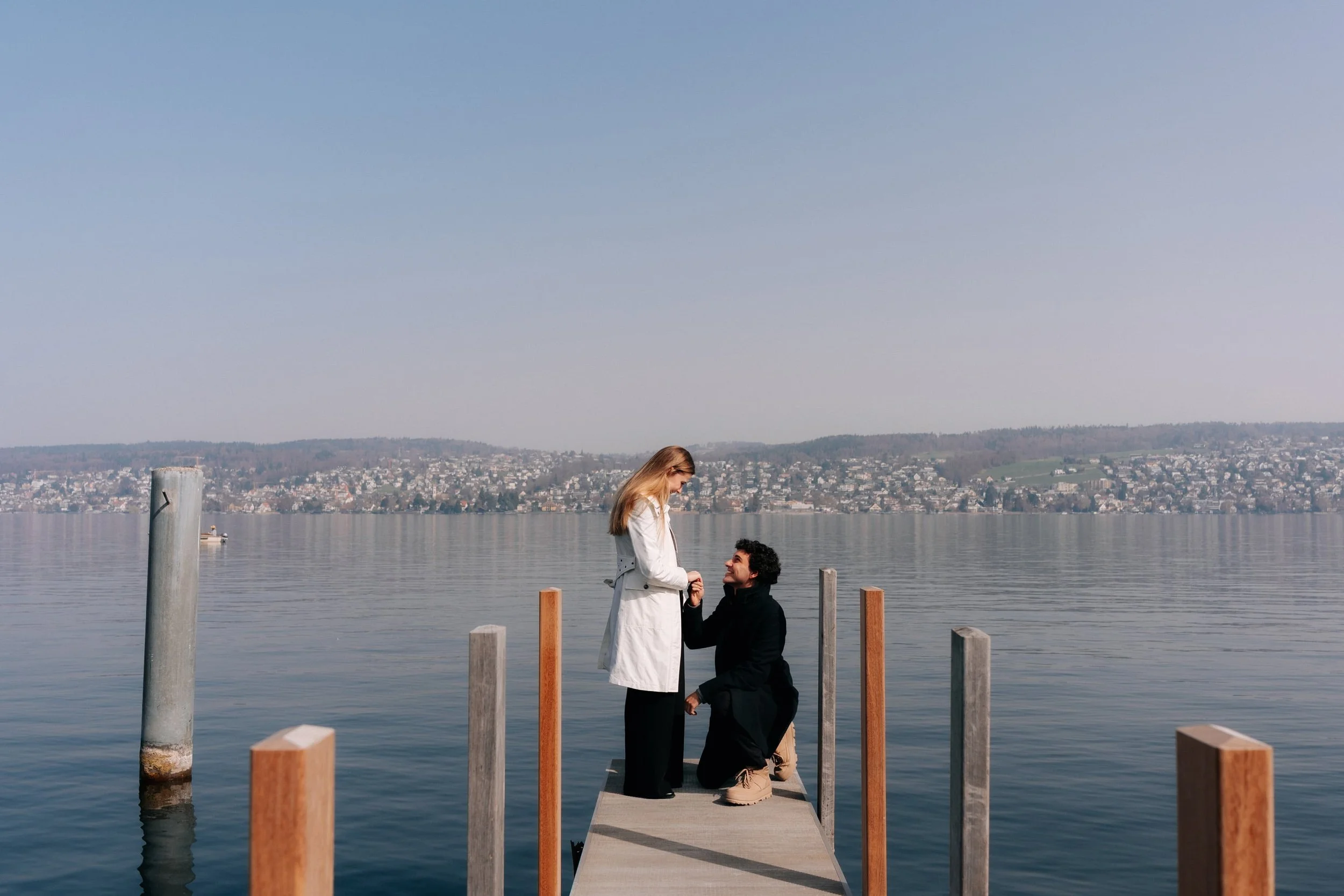 A man proposing marriage to a woman on a dock in Thalwil by Zurich lake by the water with a city and hills in the background.