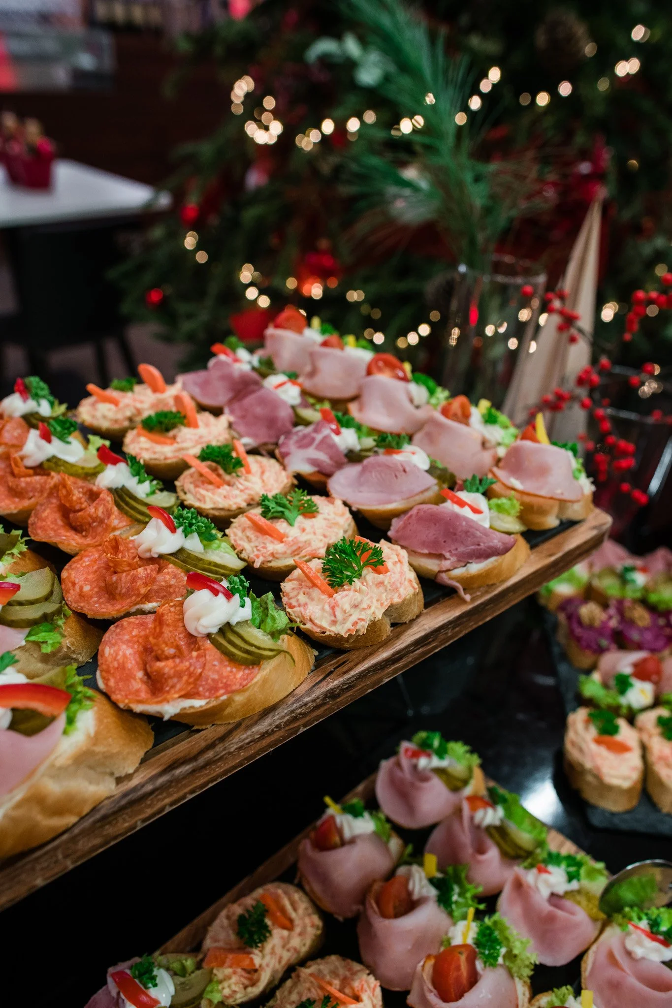 A wooden tray filled with assorted Christmas appetizers, including mini sandwiches and canapés, in front of a decorated Christmas tree with lights. Catering, culinary and food photography in Zürich, Zug, Bern, Lucerne, Basel, St. Gallen, Switzerland.
