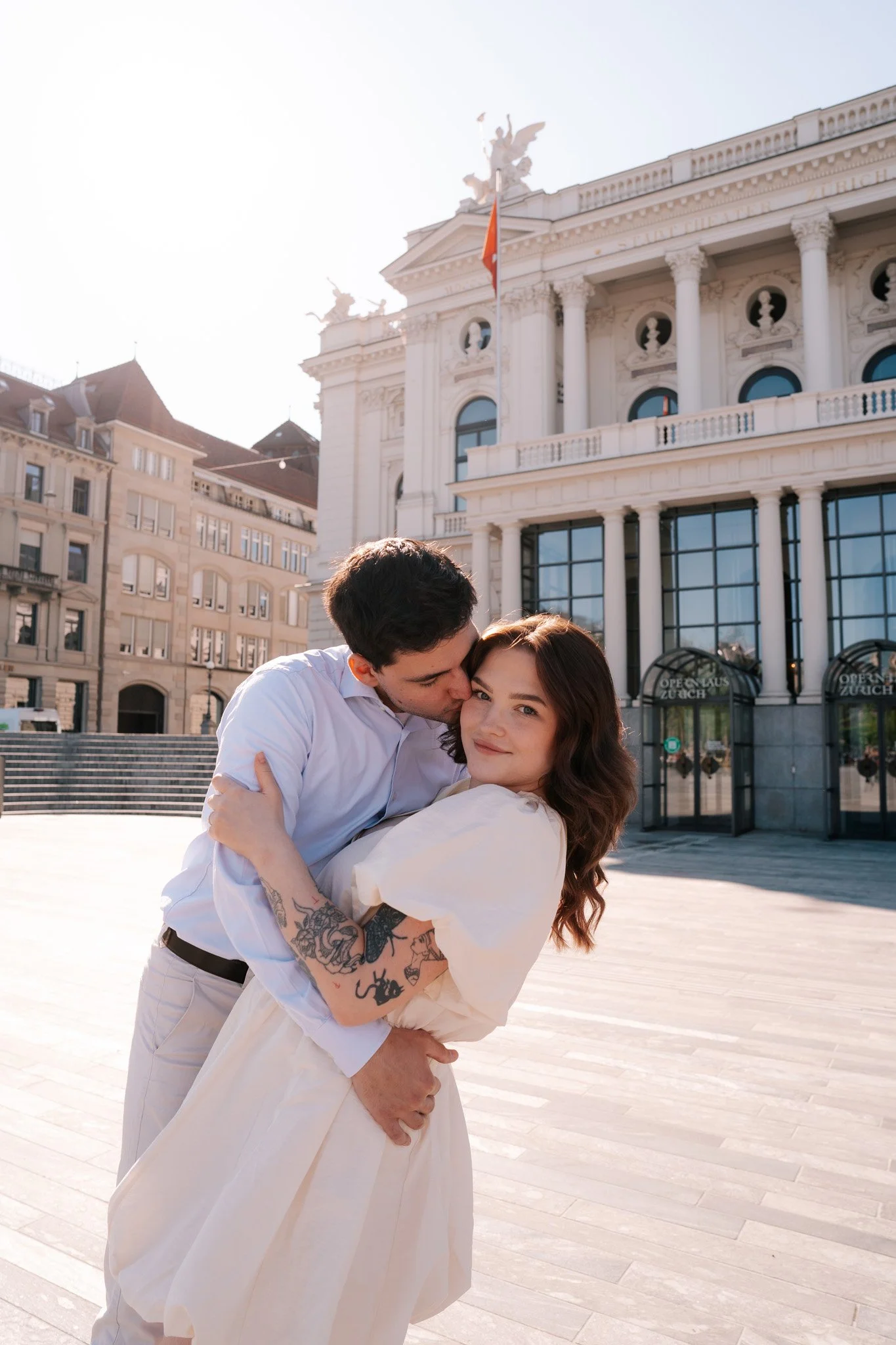 A couple embraces in front of a grand, historic building with columns and statues, on a sunny day in front of Opera in Zurich, Switzerland.