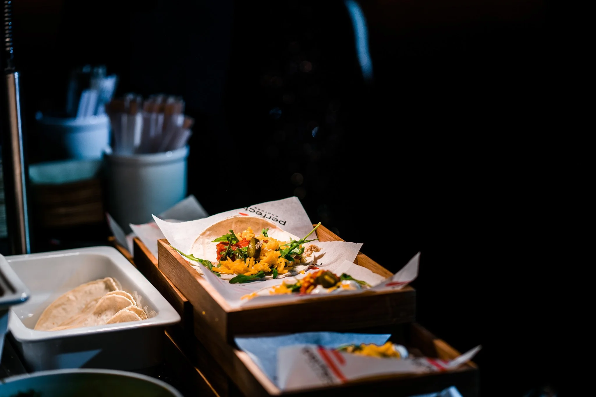 A taco with shredded cheese, vegetables, and sauce on a paper lining inside a wooden tray, with other ingredients and containers in the background. Catering and food photography in Zürich, Zug, Bern, Lucerne, Basel, St. Gallen and across Switzerland.