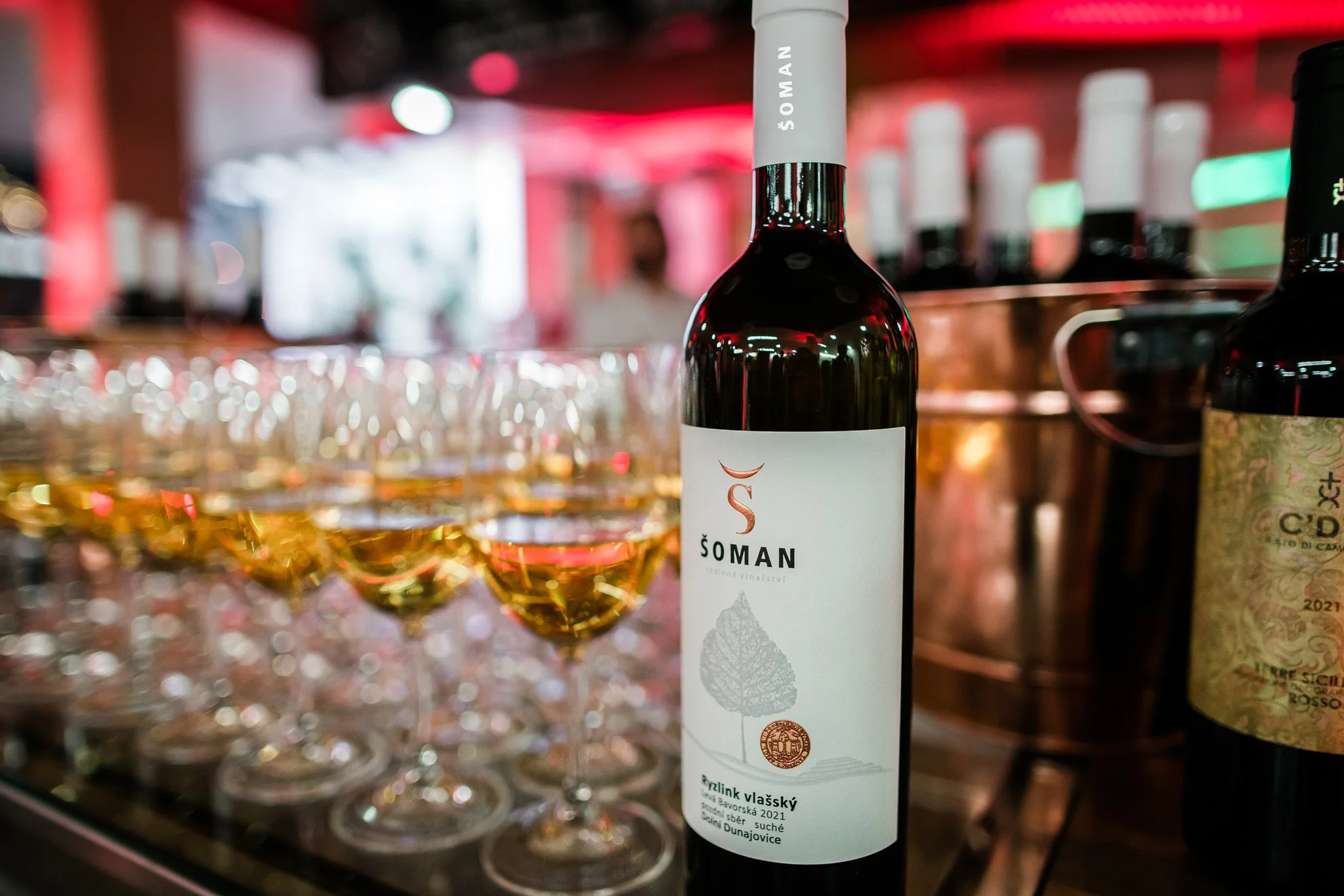 A bottle of red wine labeled 'Soman' standing on a counter with several filled wine glasses in the background and other bottles nearby, in a dimly lit setting with red and green lighting. Wine photography in Zürich, Switzerland.