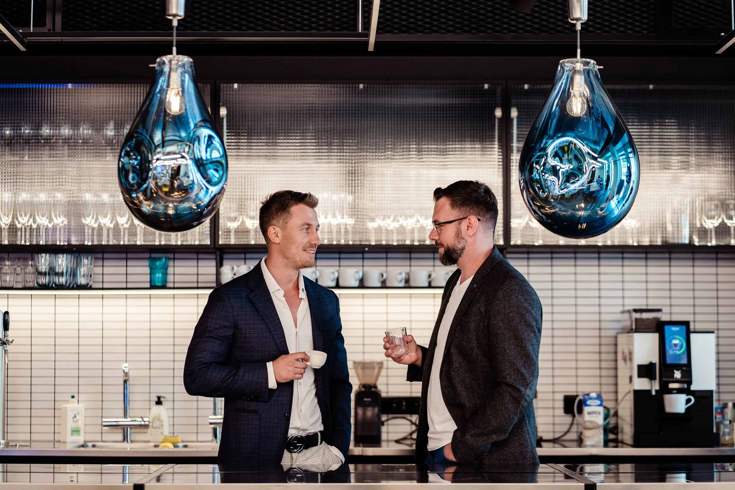 Two men in suits holding coffee cups and glasses of water, standing and talking in a modern coffee corner in the office working. Modern professional corporate branding photography in Zürich, Zug, Bern, Lucerne, Basel, St. Gallen and Switzerland.