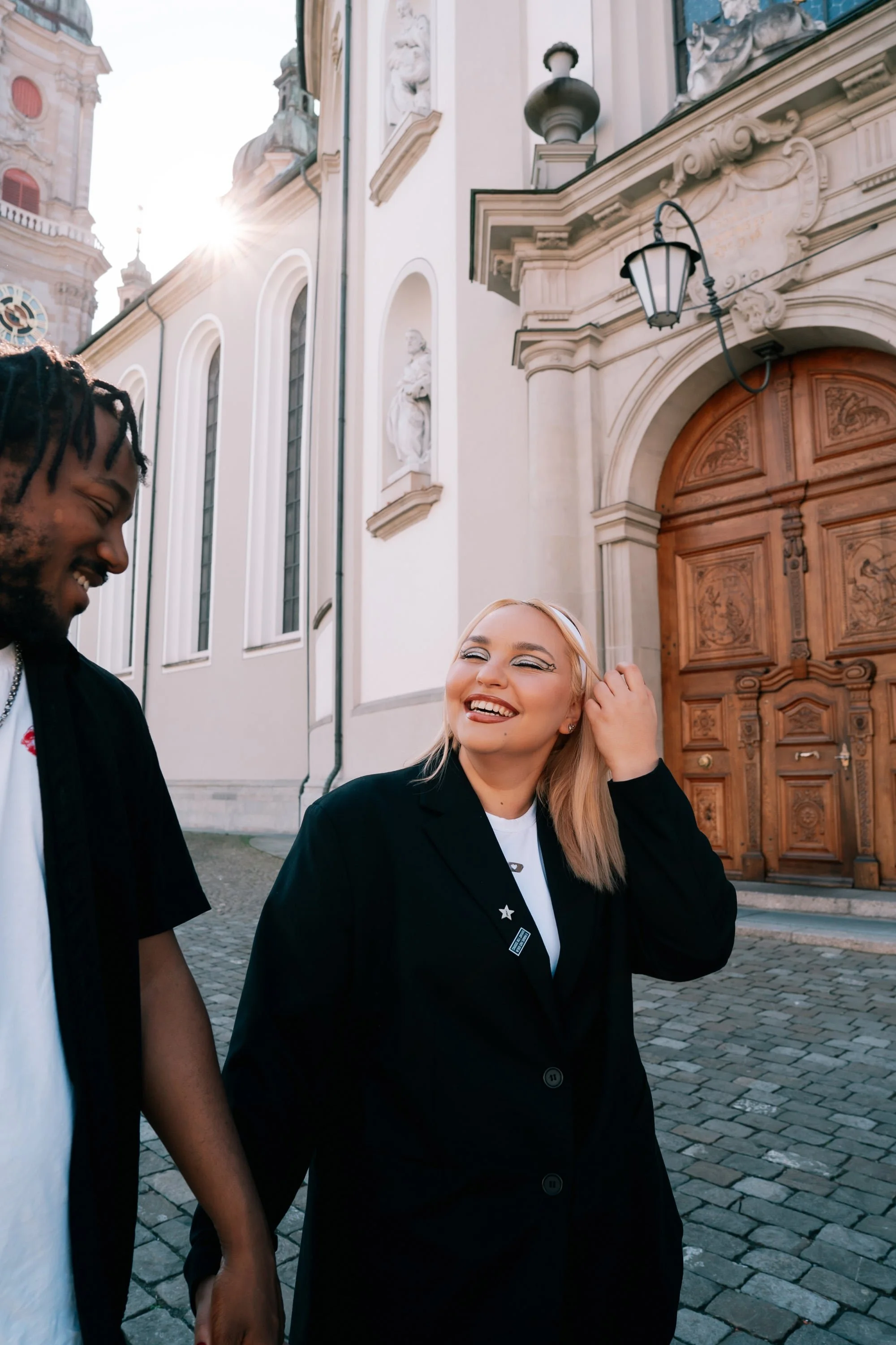 Two friends, a man and a woman, walking together outside a historic building with intricate wooden doors and sculptures, smiling and enjoying each other's company during the daytime.