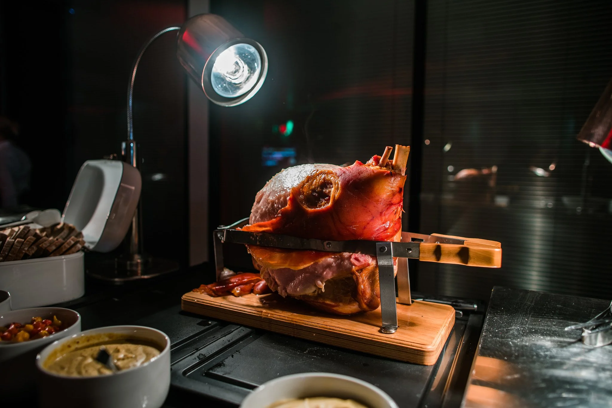 A cooked ham on a carving stand, illuminated by a focused lamp, with bowls of side dishes nearby in a dimly lit setting. Catering and food photography in Zürich, Zug, Bern, Lucerne, Basel, St. Gallen and across Switzerland.