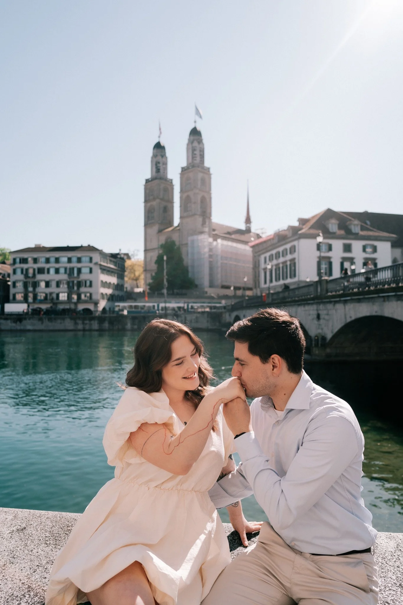 A woman and a man sitting by a river, holding hands and touching foreheads, with a historic church and bridge in the background on a sunny day in Zurich Switzerland.