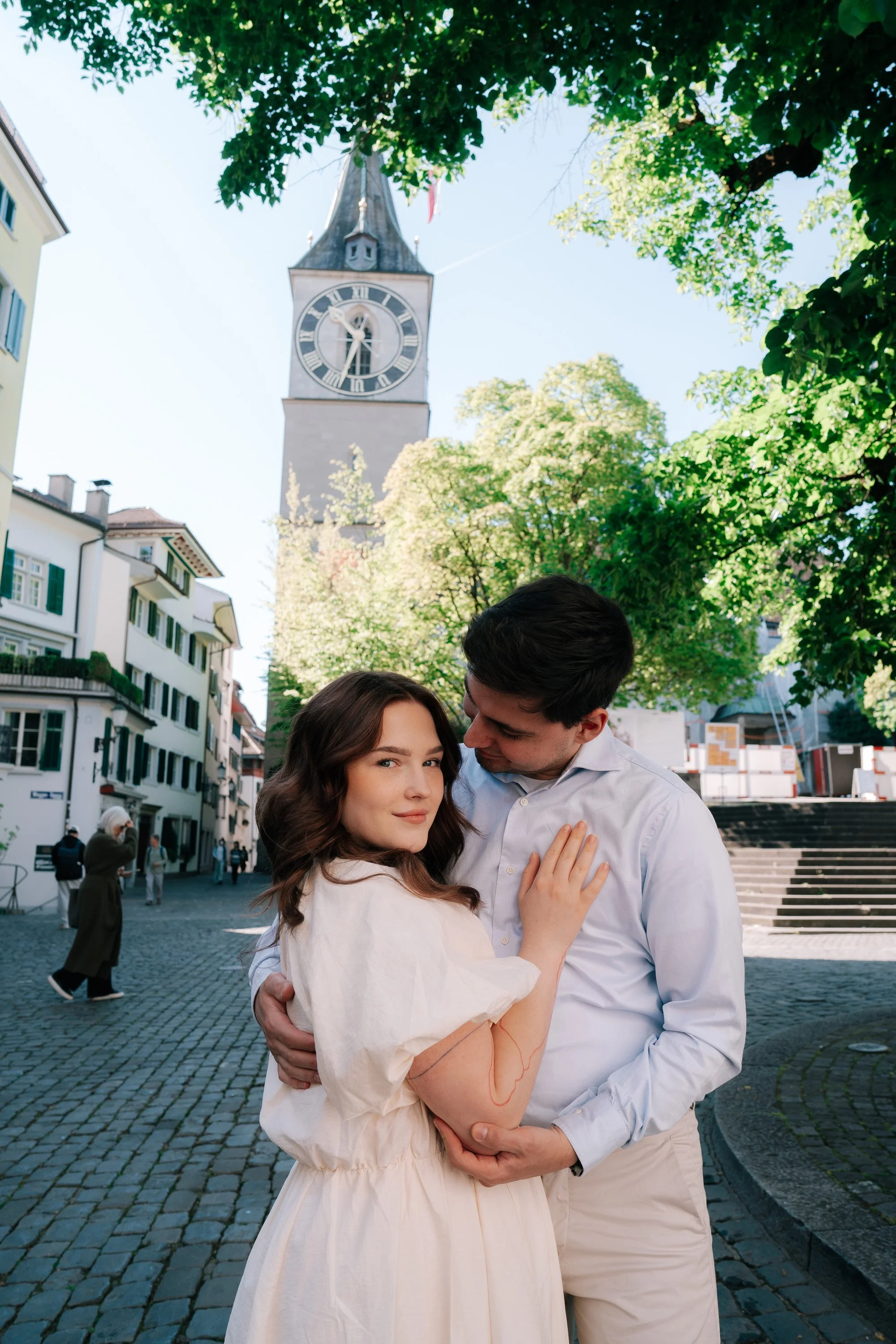 A couple embracing on a cobblestone street with a clock tower in the background, surrounded by green trees and buildings in Zurich Switzerland.