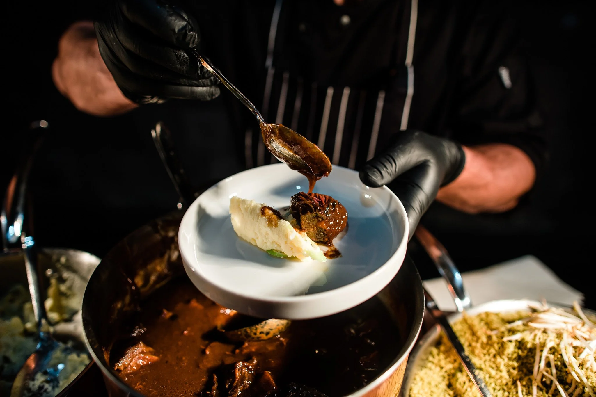 Person wearing black gloves serving a scoop of meat stew with mashed potatoes on a white plate. There are other dishes in the background. Catering, culinary and food photography in Zürich, Zug, Bern, Lucerne, Basel, St. Gallen and across Switzerland.
