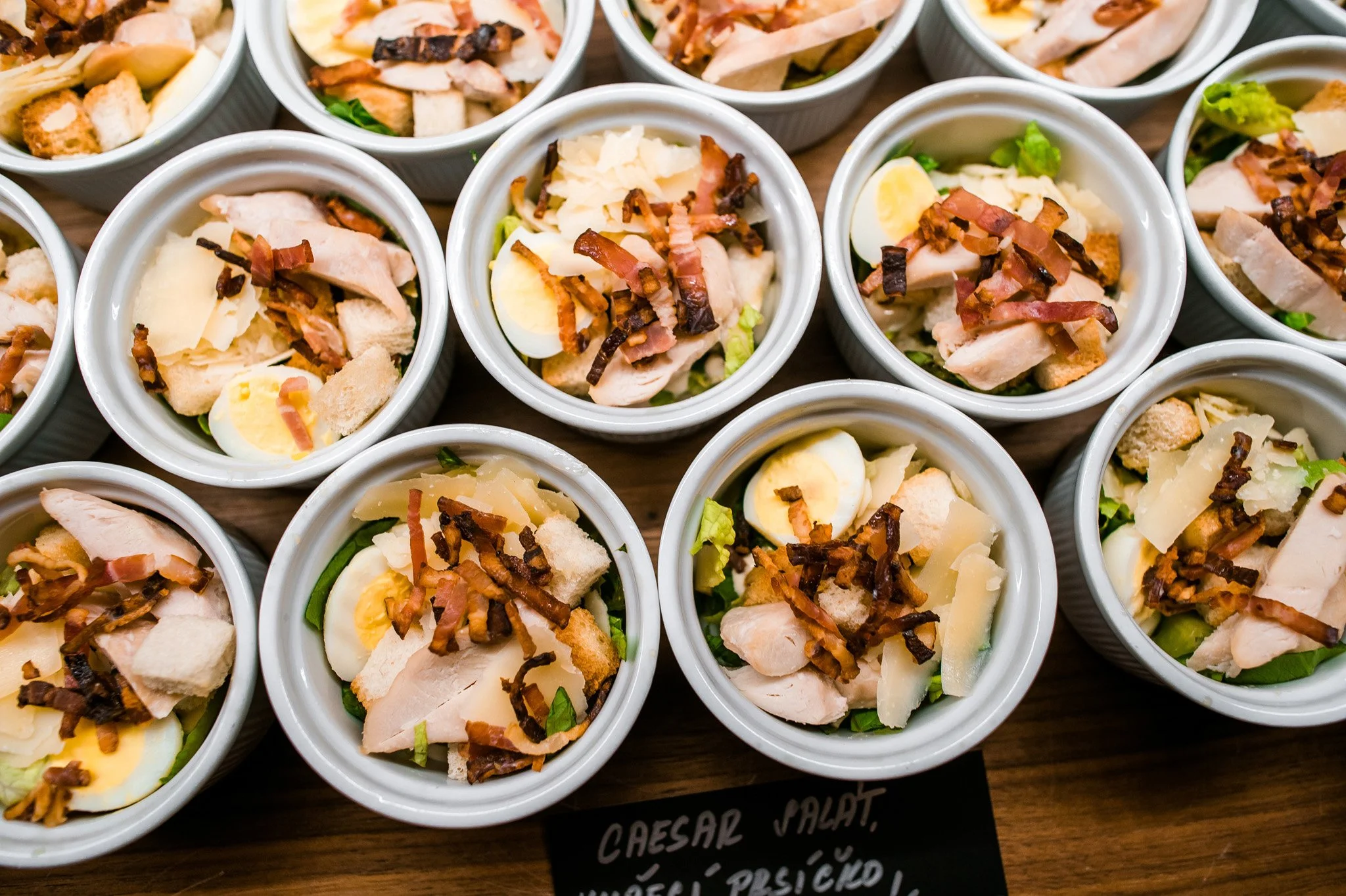 Small bowls of Caesar salad topped with grilled chicken, bacon bits, Parmesan cheese, croutons, and boiled eggs on a wooden surface. Catering and food photography in Zürich, Zug, Bern, Lucerne, Basel, St. Gallen and across Switzerland.