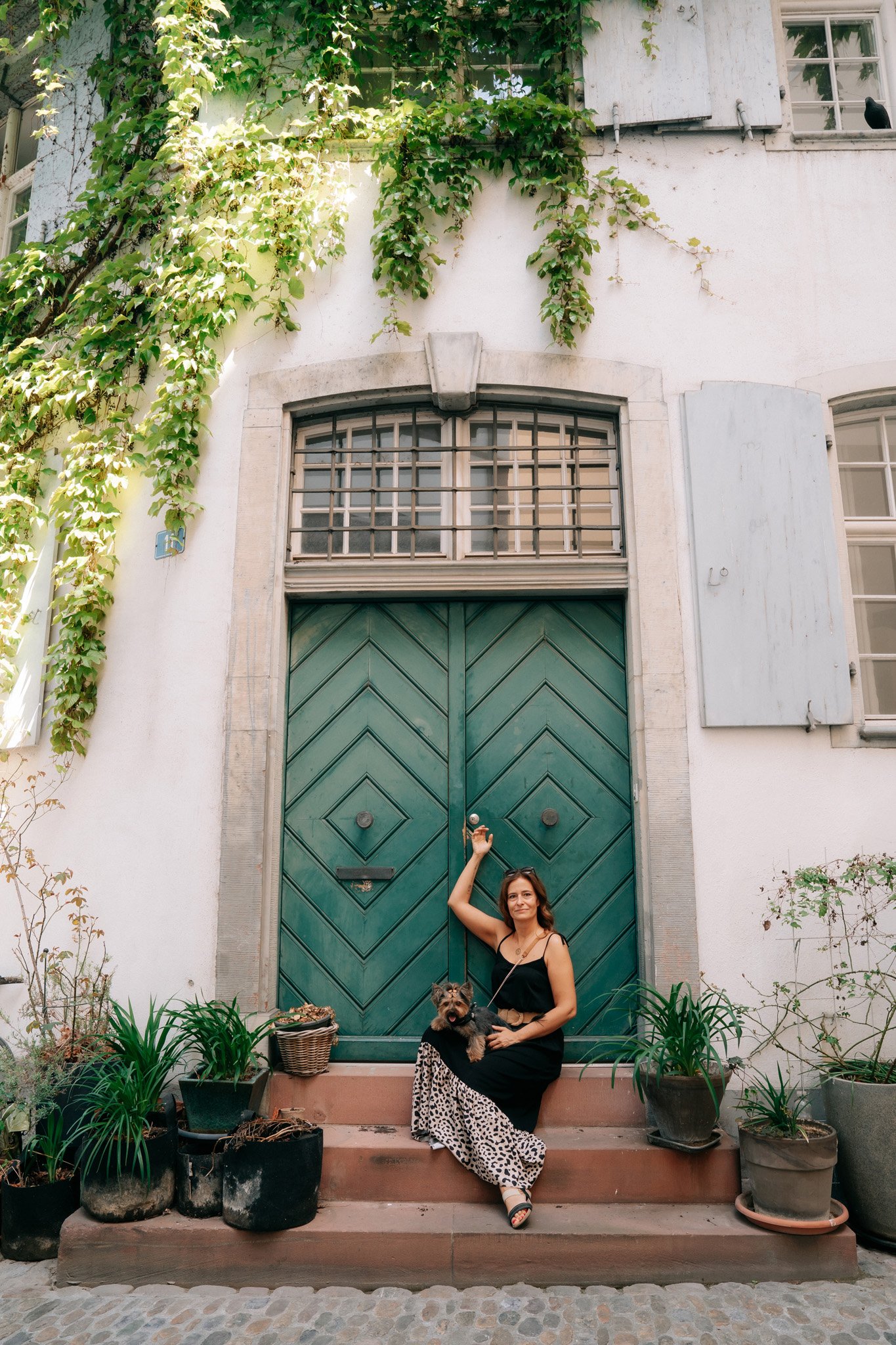 Photoshoot of solo traveler sitting on steps with small dog in front of large green door in Basel, Switzerland, surrounded by potted plants, old building with white walls and windows.