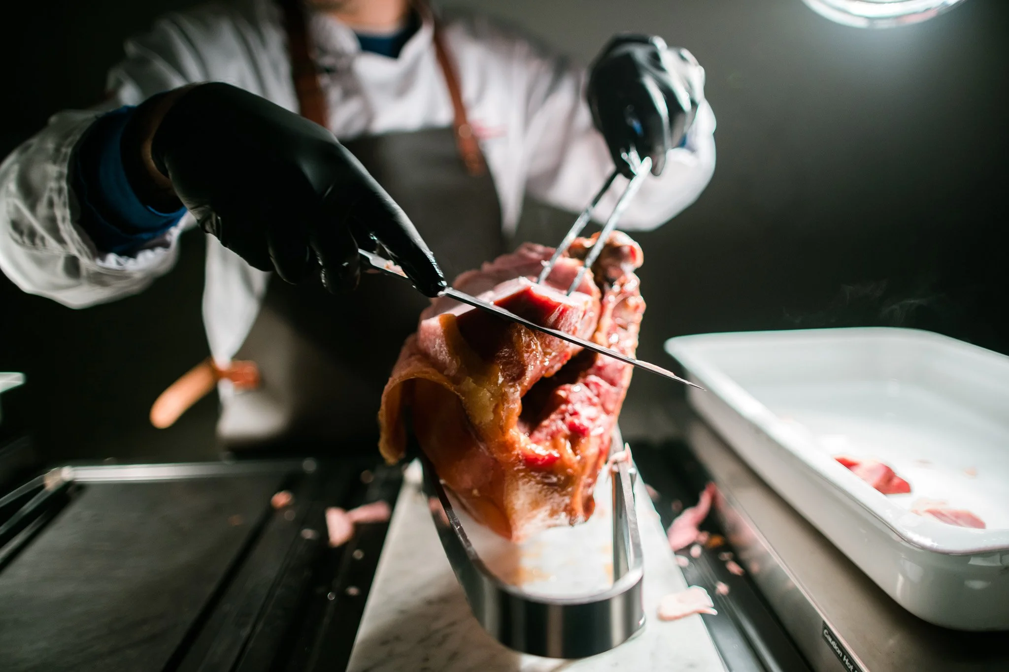 Chef in black gloves carving a large cut of meat with a knife and fork in a professional kitchen. Catering and food photography in Zürich, Zug, Bern, Lucerne, Basel, St. Gallen and across Switzerland.