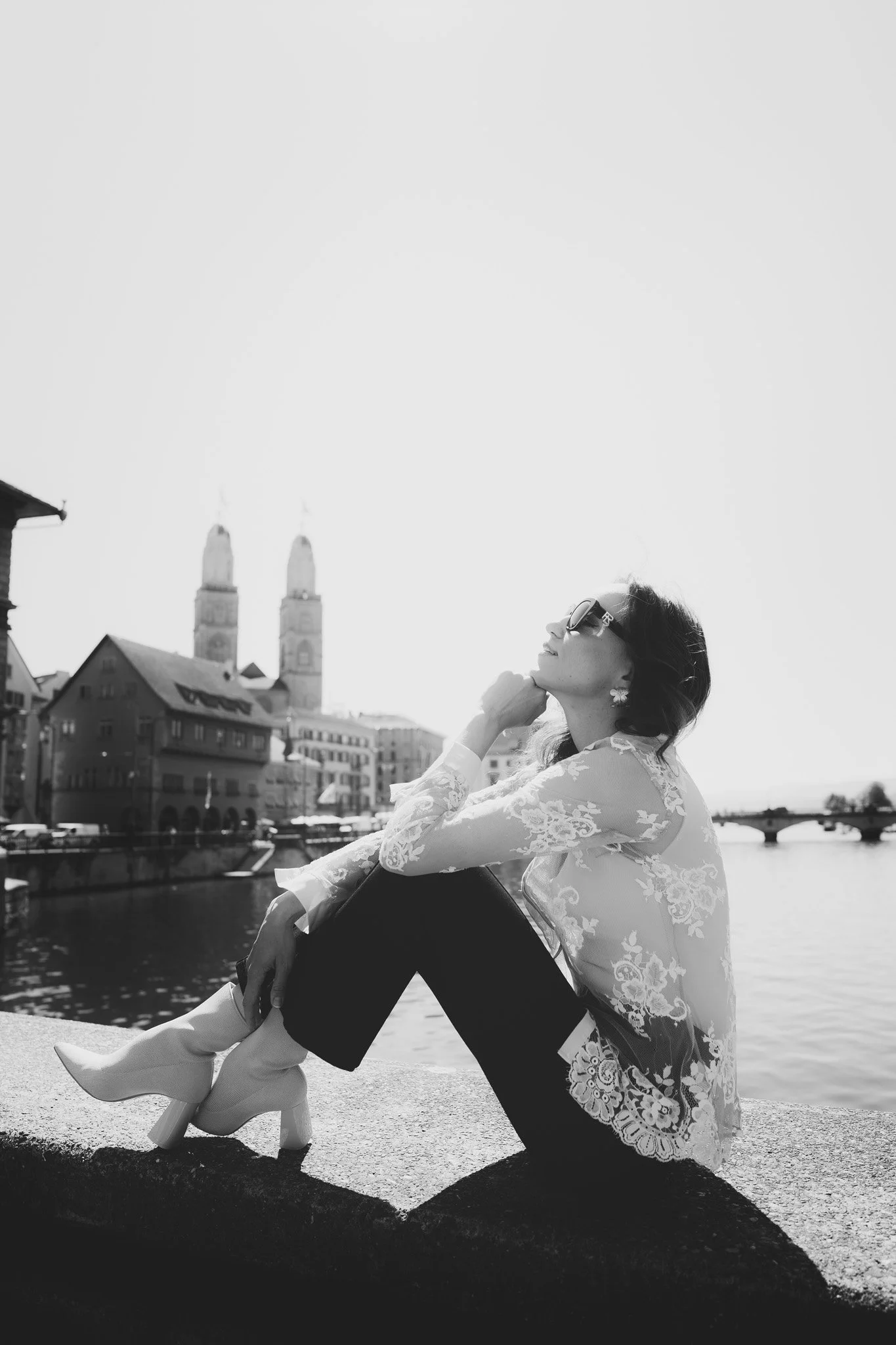 A woman sitting outdoors by a river with historic buildings and a church of Zurich Switzerland in the background. She is wearing sunglasses, lace top, and high-heeled boots, relaxing with her eyes closed.