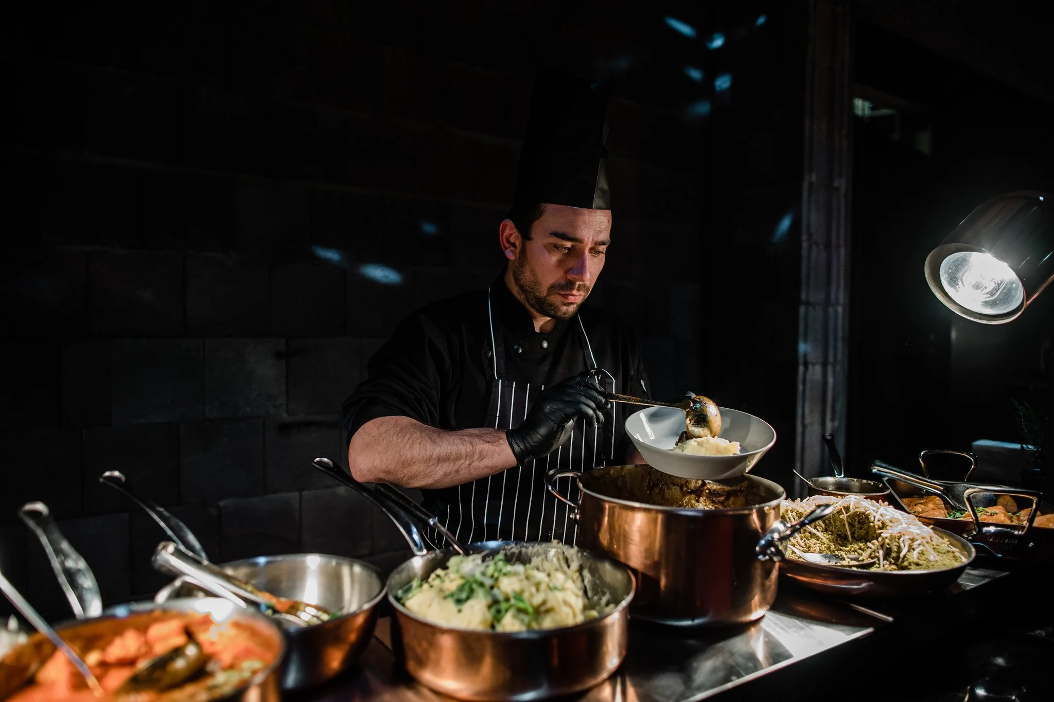A male chef in a black uniform and hat ladles food into a white dish at a buffet station with various prepared dishes, in a dimly lit kitchen or restaurant setting. Catering and food photography in Zürich, Zug, Bern, Lucerne, Basel, St. Gallen.
