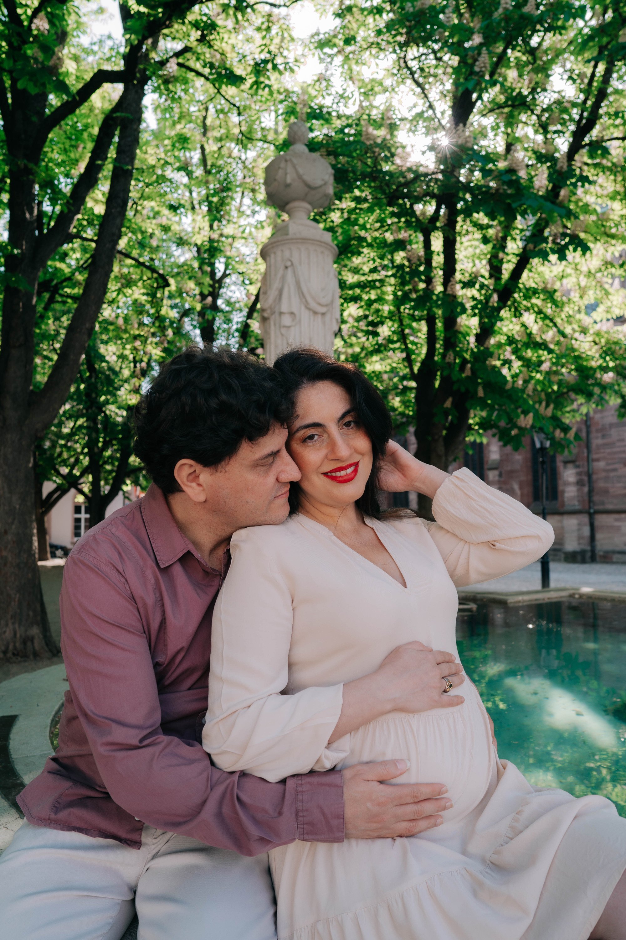 Couple sitting and smiling during Basel maternity photoshoot by the fountain.
