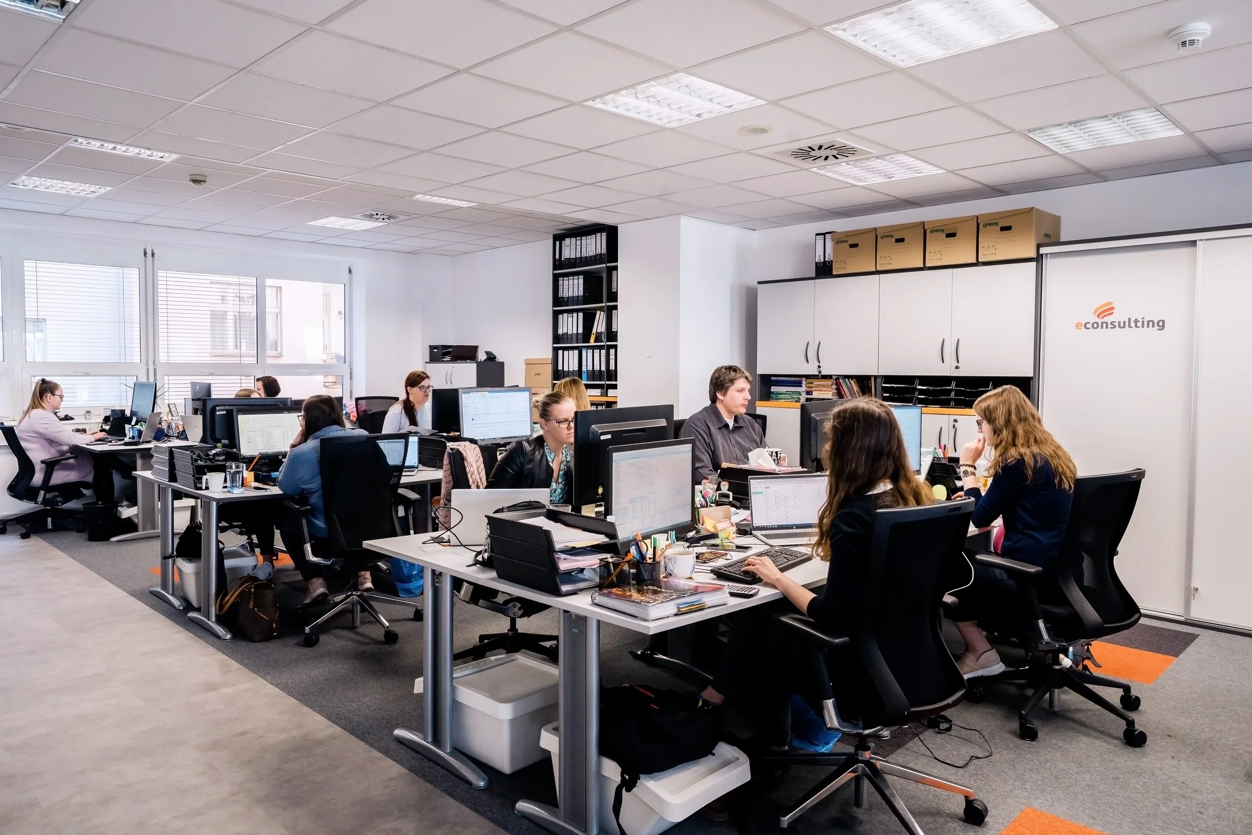 Office workspace with employees working at desks with multiple computer monitors, in a bright room with windows and white cabinets, enclosed by glass walls. Office photography in Zürich, Zug, Bern, Lucerne, Basel, St. Gallen and across Switzerland.