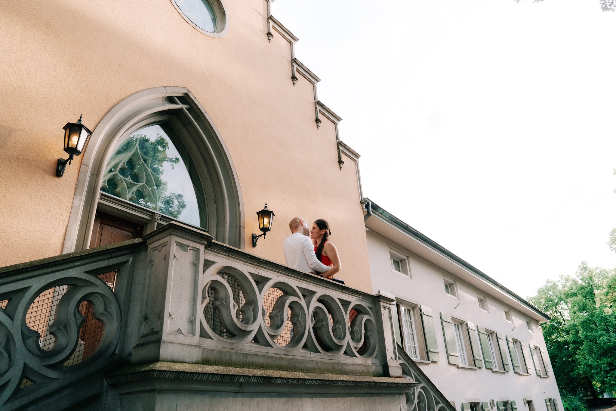 A couple standing on a balcony of a European-style building, looking at each other and smiling. The building has a pinkish stucco exterior, large arched window with spider web-like decoration, and black lantern-style light fixtures. There are green t