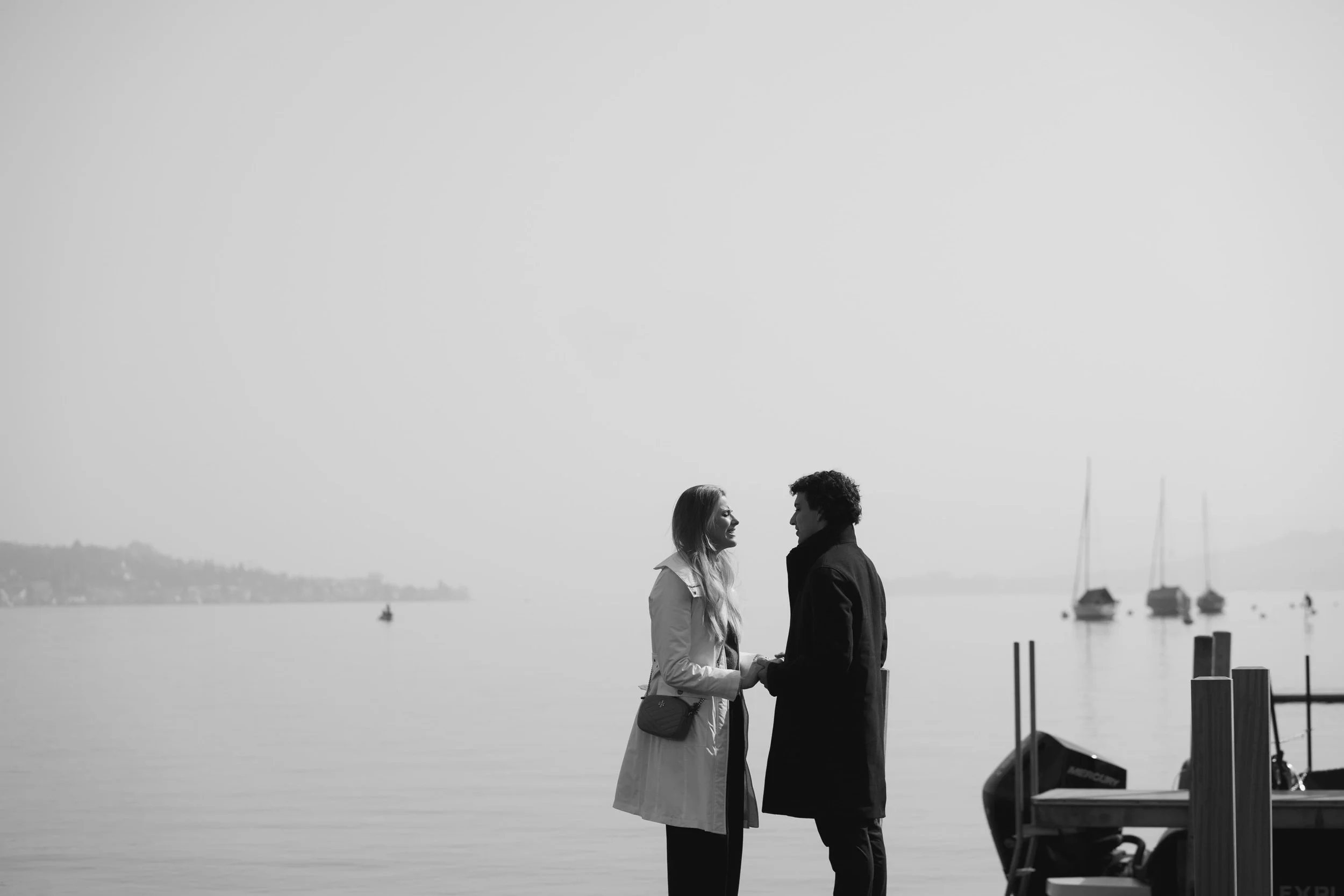 A black-and-white photo of a couple holding hands by the water with sailboats in the background during a romantic proposal in Thalwil in Switzerland near a lake.