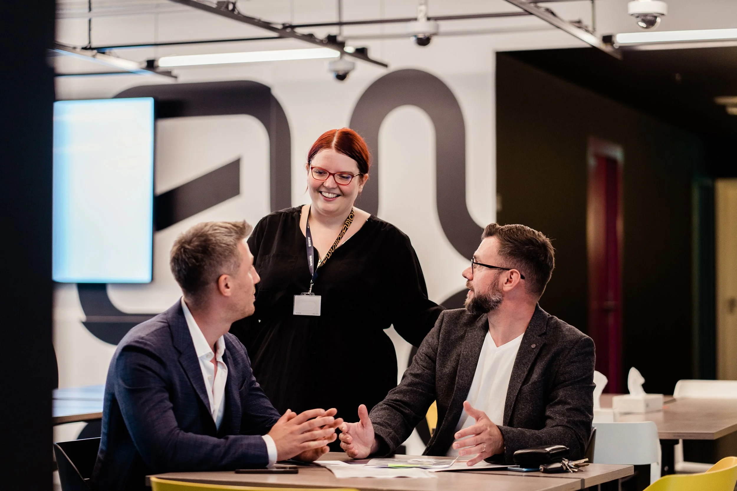 Two men having a discussion while seated at a table, with a woman standing and smiling between them, in a modern conference room or workspace. Modern employer branding photography in Zürich, Zug, Bern, Lucerne, Basel, St. Gallen - across Switzerland.