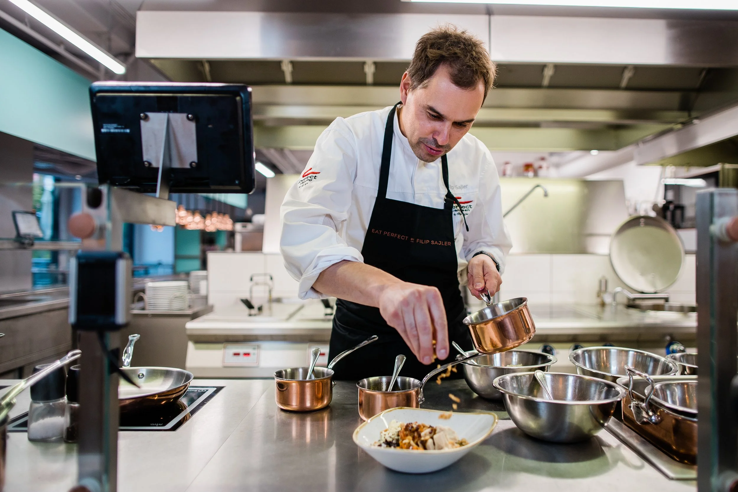 A chef in a professional kitchen is carefully plating a dish with small pots and bowls around him, wearing a white shirt and black apron. Personal branding and fine dining restaurant photography in Zürich, Zug, Bern, Lucerne, Basel, St. Gallen and ac