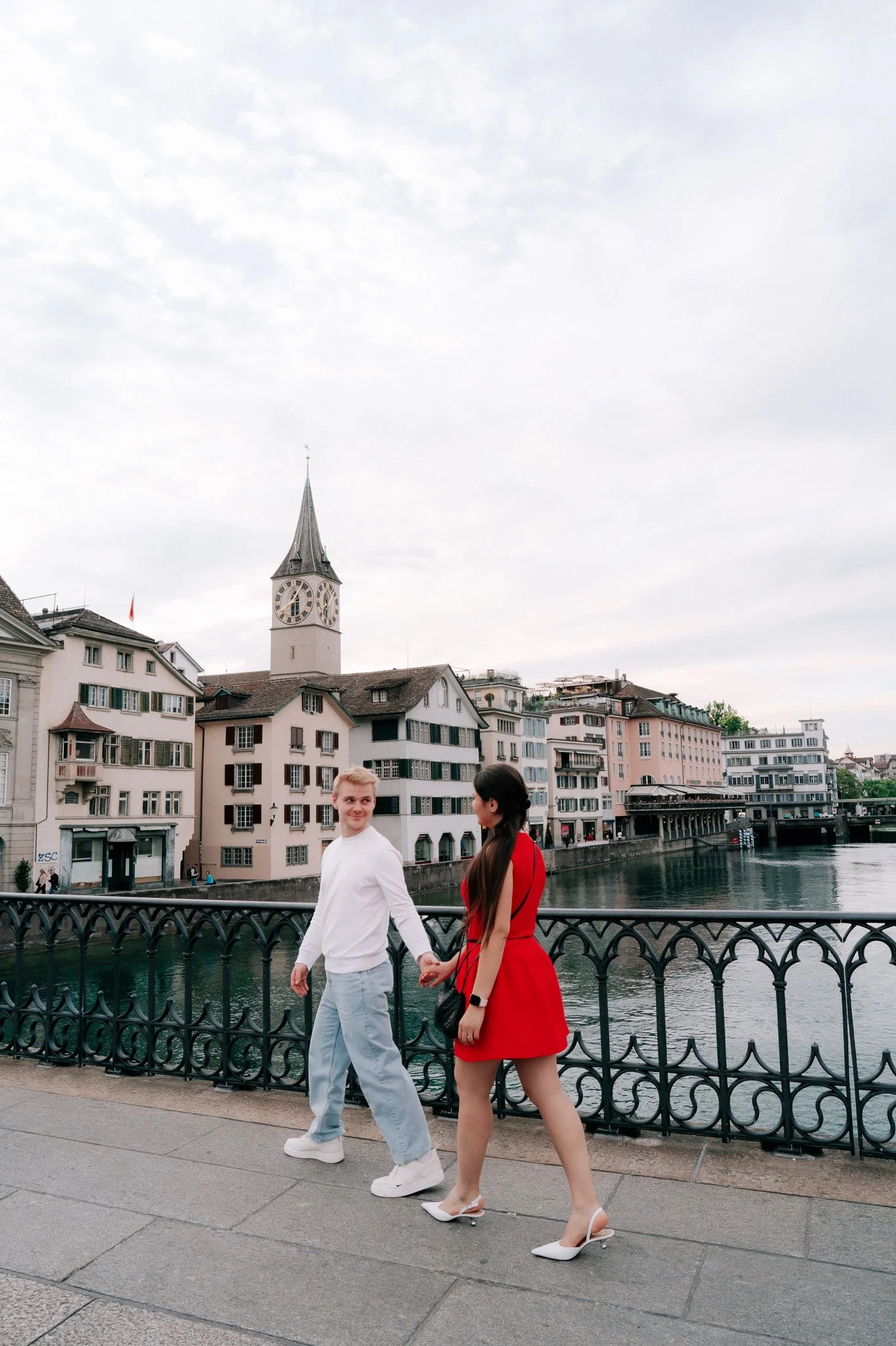 A couple holding hands and walking along a riverfront with city buildings and a clock tower in the background.