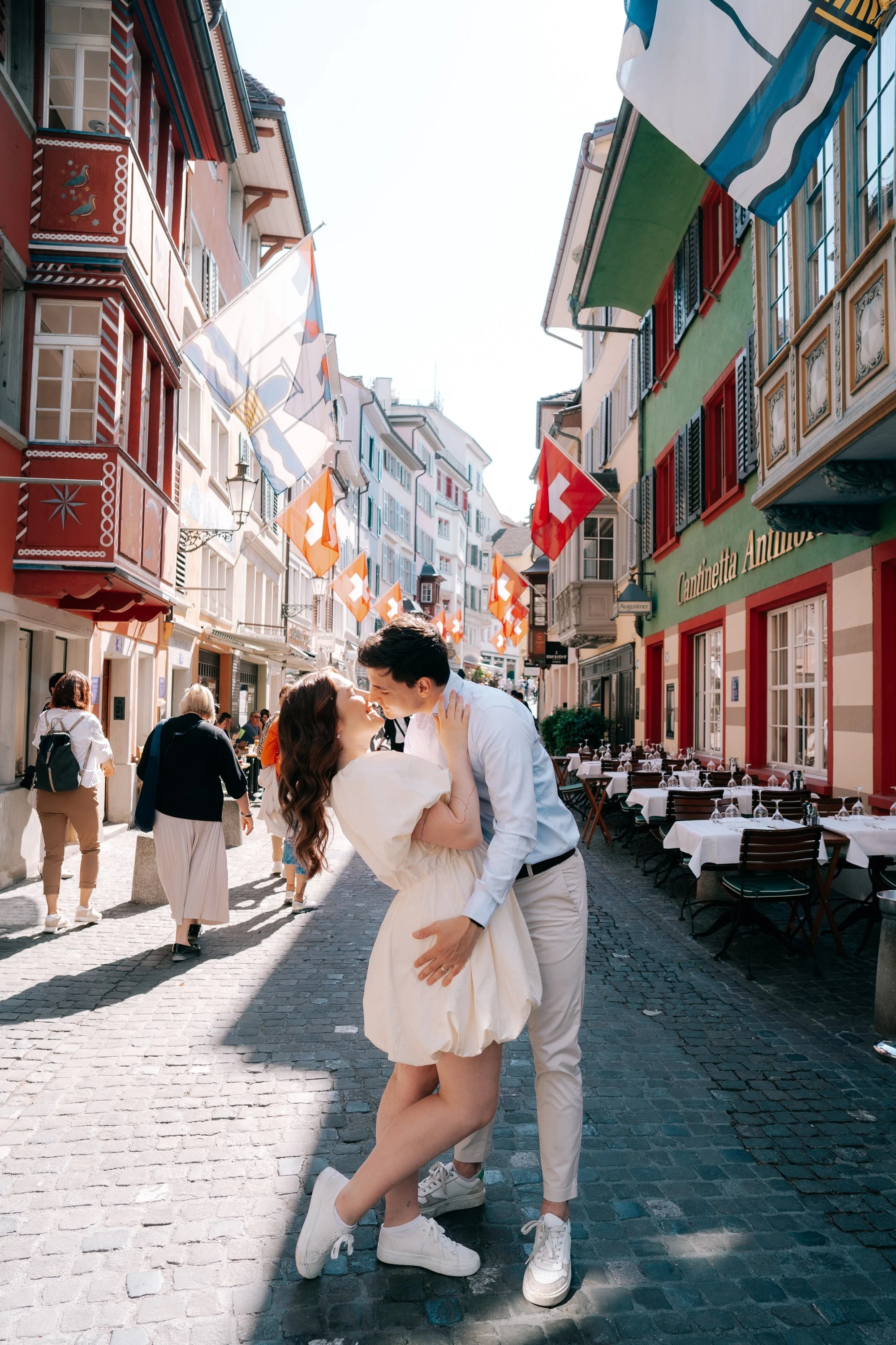 A couple kissing on a cobblestone street in Zurich Switzerland, with colorful buildings and Swiss flags hanging overhead.