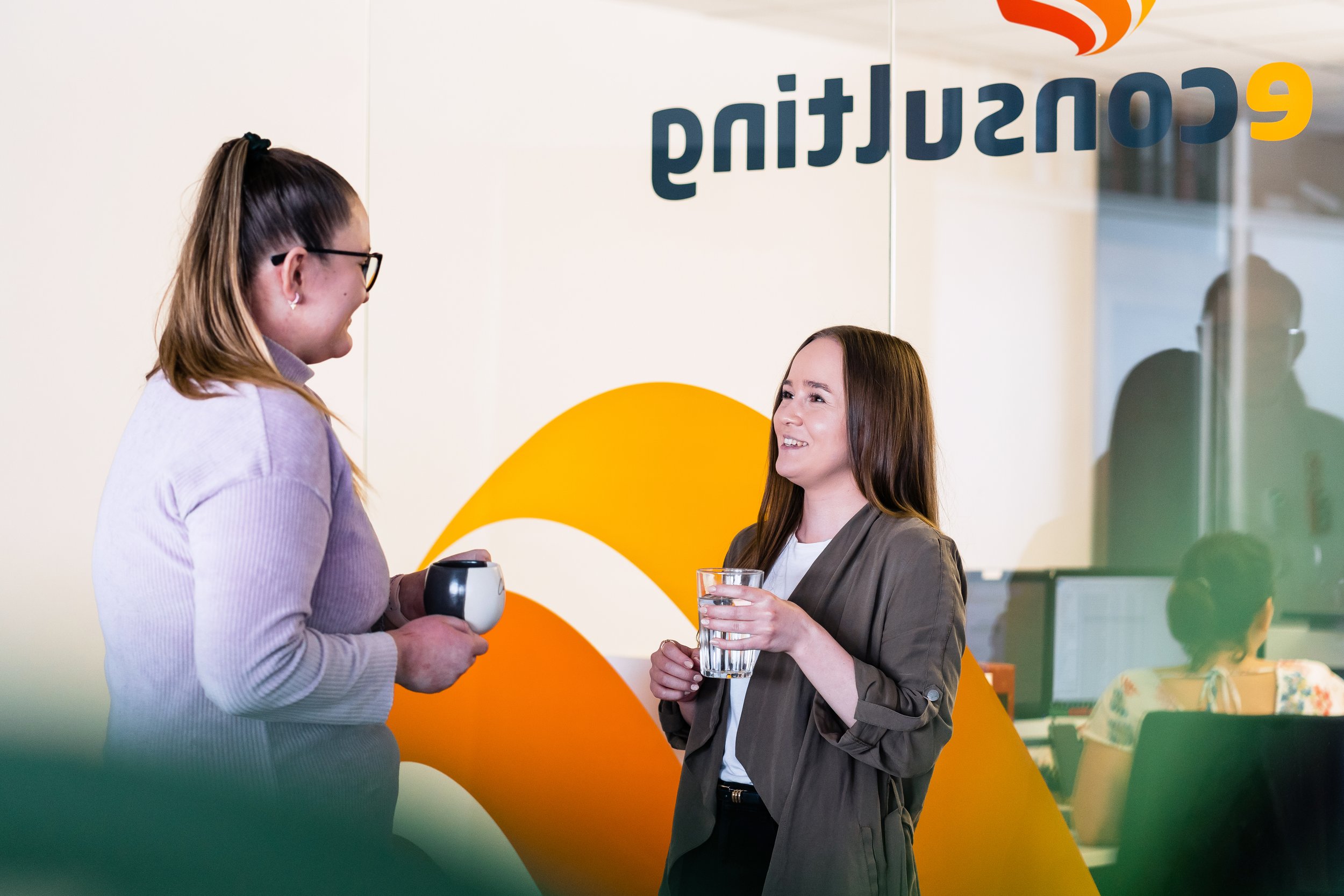 Two women engaged in conversation in an office, one holding a coffee mug and the other holding a glass of water, with a colorful sign in the background. Corporate photography in Zürich, Zug, Bern, Lucerne, Basel, St. Gallen and across Switzerland.