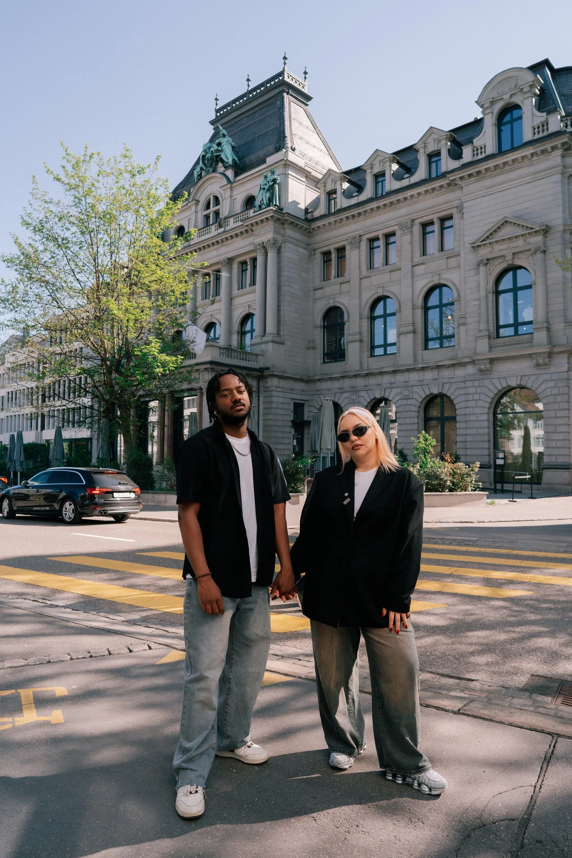 Couple fashionably dressed holding hands stand on a crosswalk in front of a historic building with ornate architecture in an urban street in St. Gallen Switzerland.
