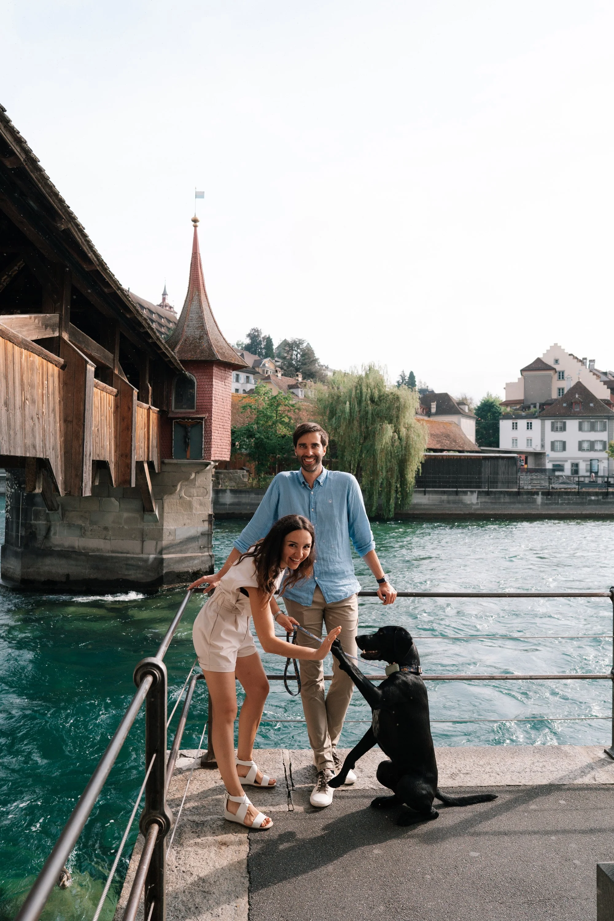 A smiling couple with a black dog on a leash giving high five, standing on a dock by a body of water with buildings in Lucerne Switzerland and a small church in the background.