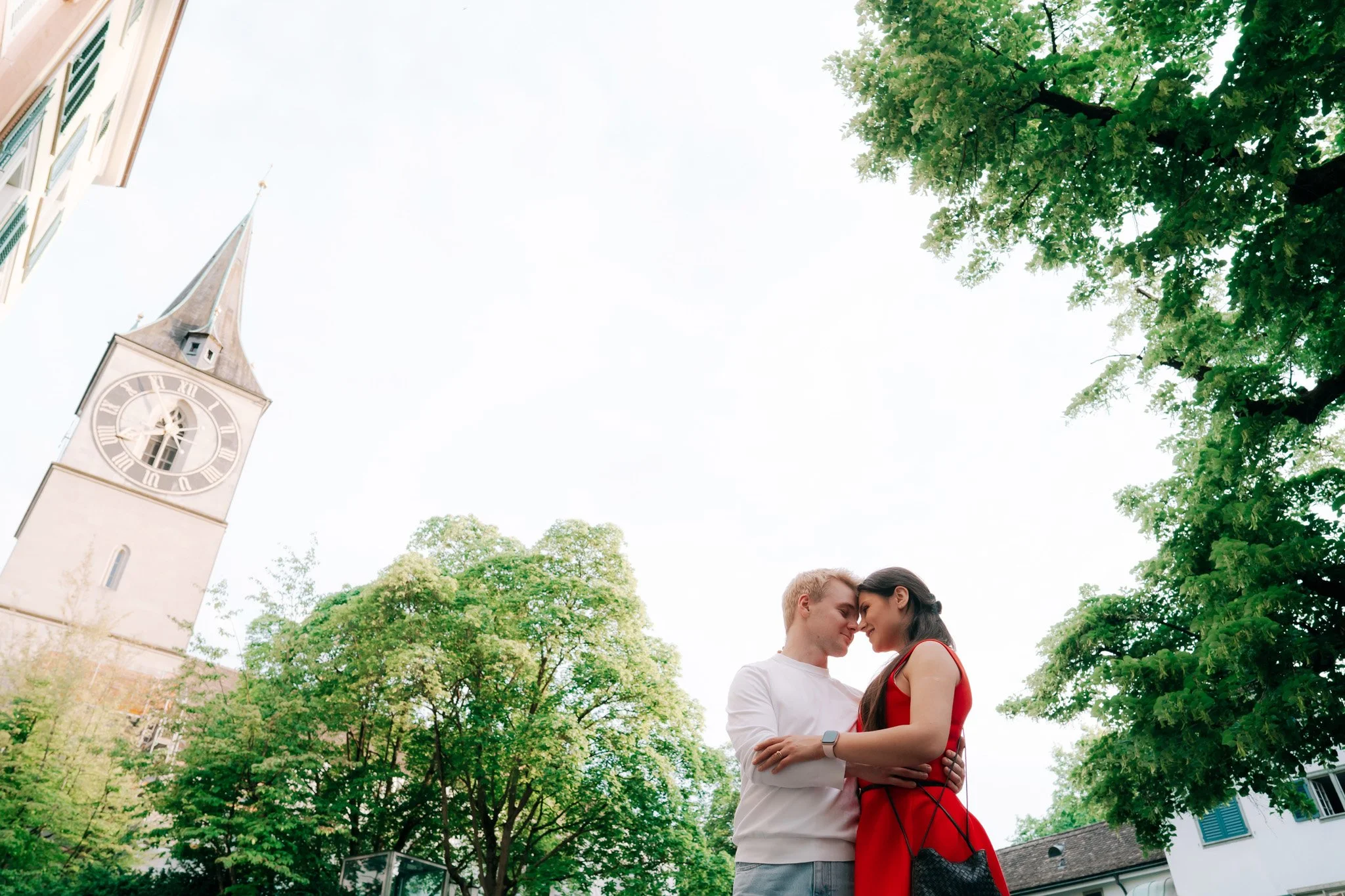 A couple with foreheads touching, smiling, embracing outdoors with greenery and a clock tower in the background in historic city centre of Zurich Switzerland.
