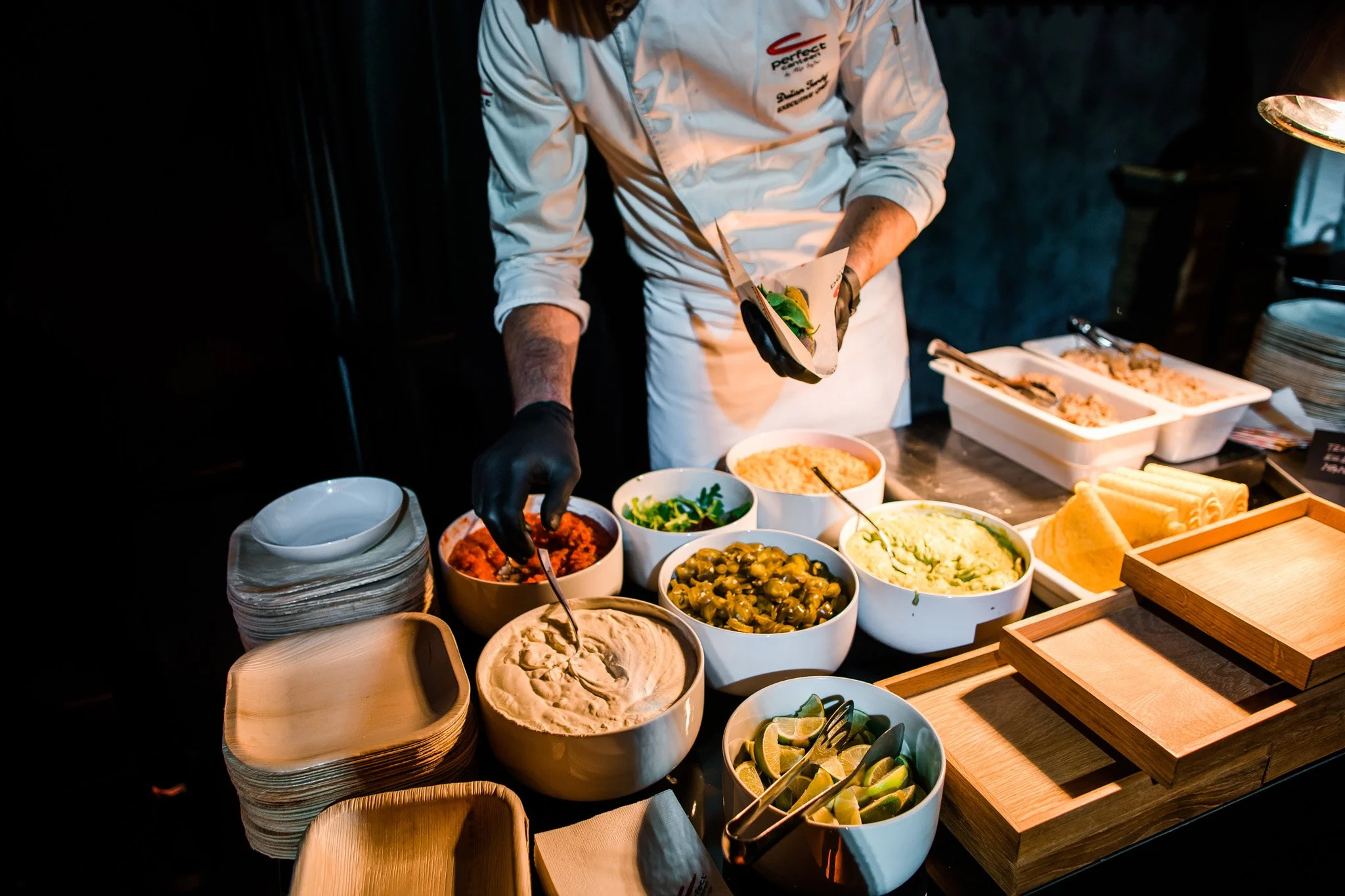 A chef in a white uniform and black gloves prepares a taco at a buffet station with bowls of various toppings such as diced tomatoes, guacamole, jalapeños, shredded cheese, and lime wedges. Catering, culinary and food photography in Switzerland.
