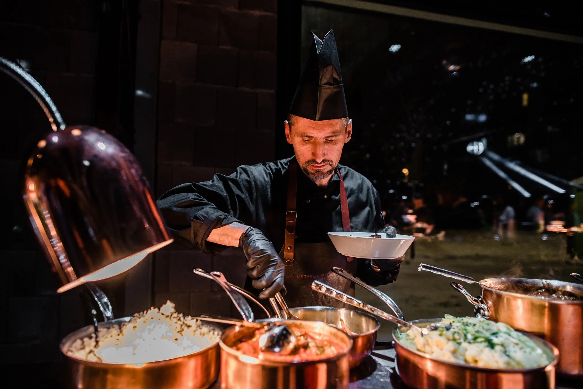 A chef in a black hat and apron serves food at an outdoor event during nighttime. Multiple copper pots of food are on a counter, with steam rising from some of them. Catering, culinary and food photography in Zürich, Zug, Bern, and across Switzerland