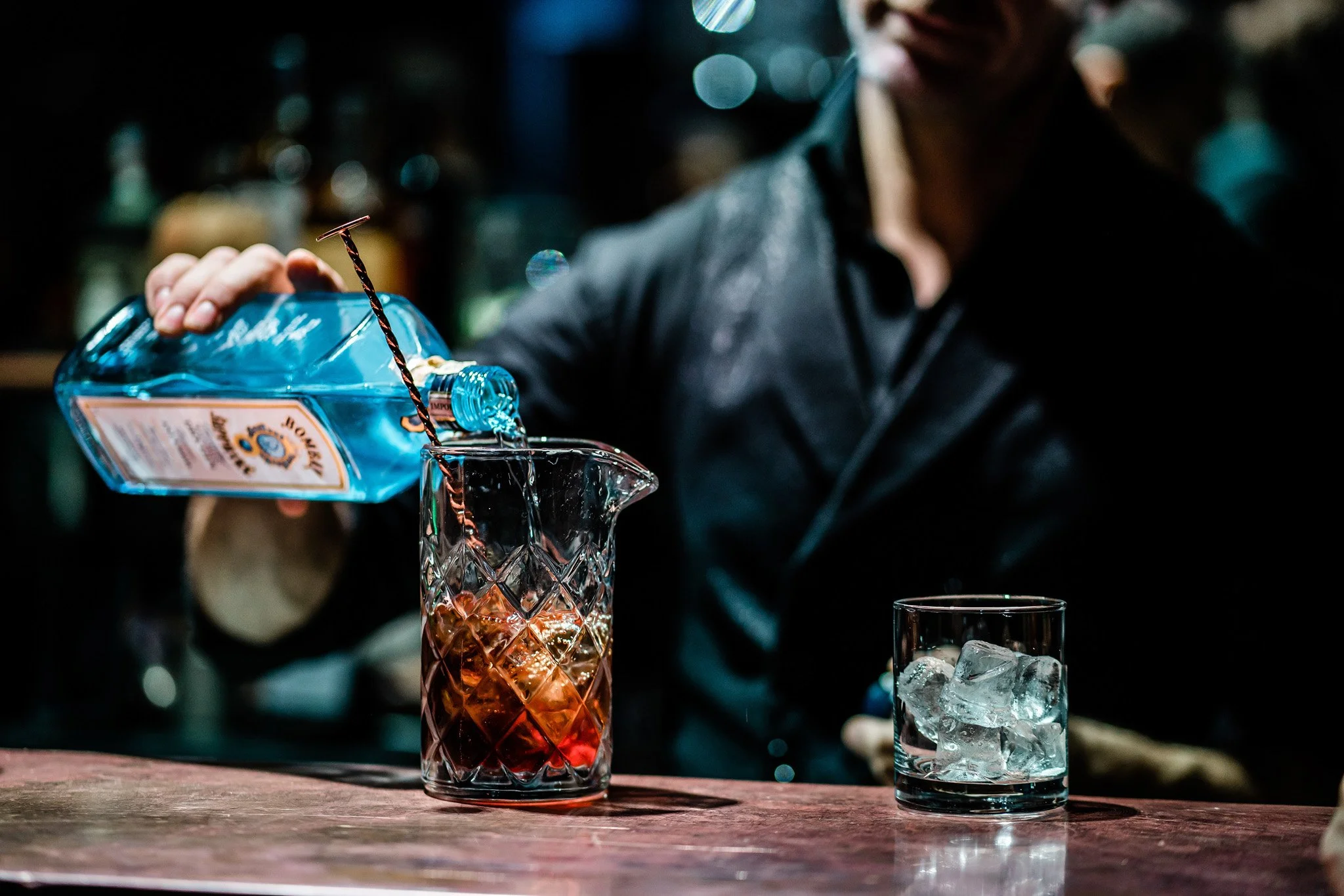 A bartender pouring a bottle of blue liquid into a glass with ice and a carved glass container behind a wooden bar. Catering alcohol cocktail bar and food photography in Zürich, Zug, Bern, Lucerne, Basel, St. Gallen and across Switzerland.
