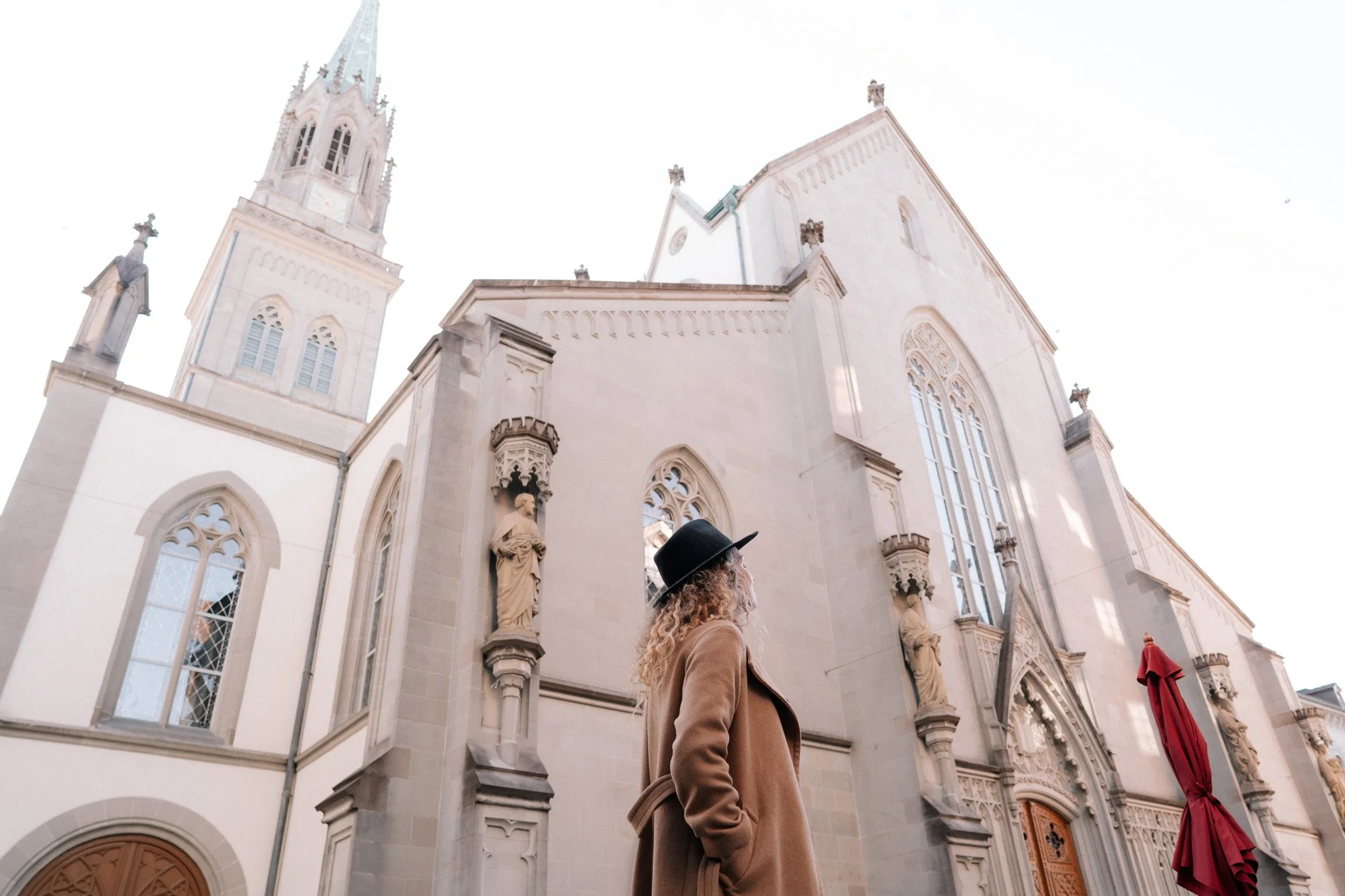 A person with curly blonde hair wearing a black hat and brown coat standing in front of a grand, white Gothic-style church in St. Gallen, Switzerland, with tall towers and detailed sculptures.