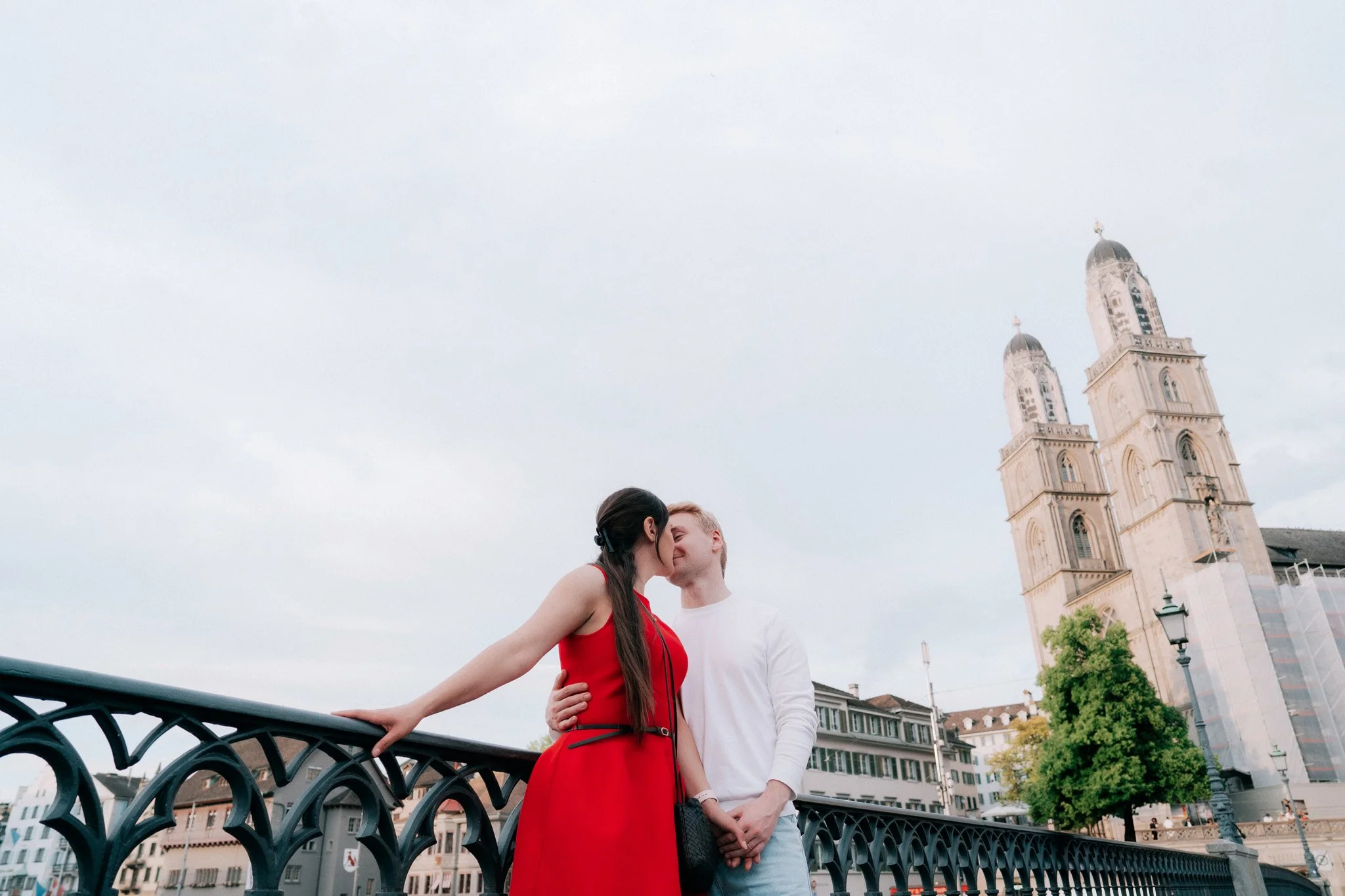 A couple sharing a kiss on a bridge in front of a large historic building with twin towers of Grossmunster and a cloudy sky.