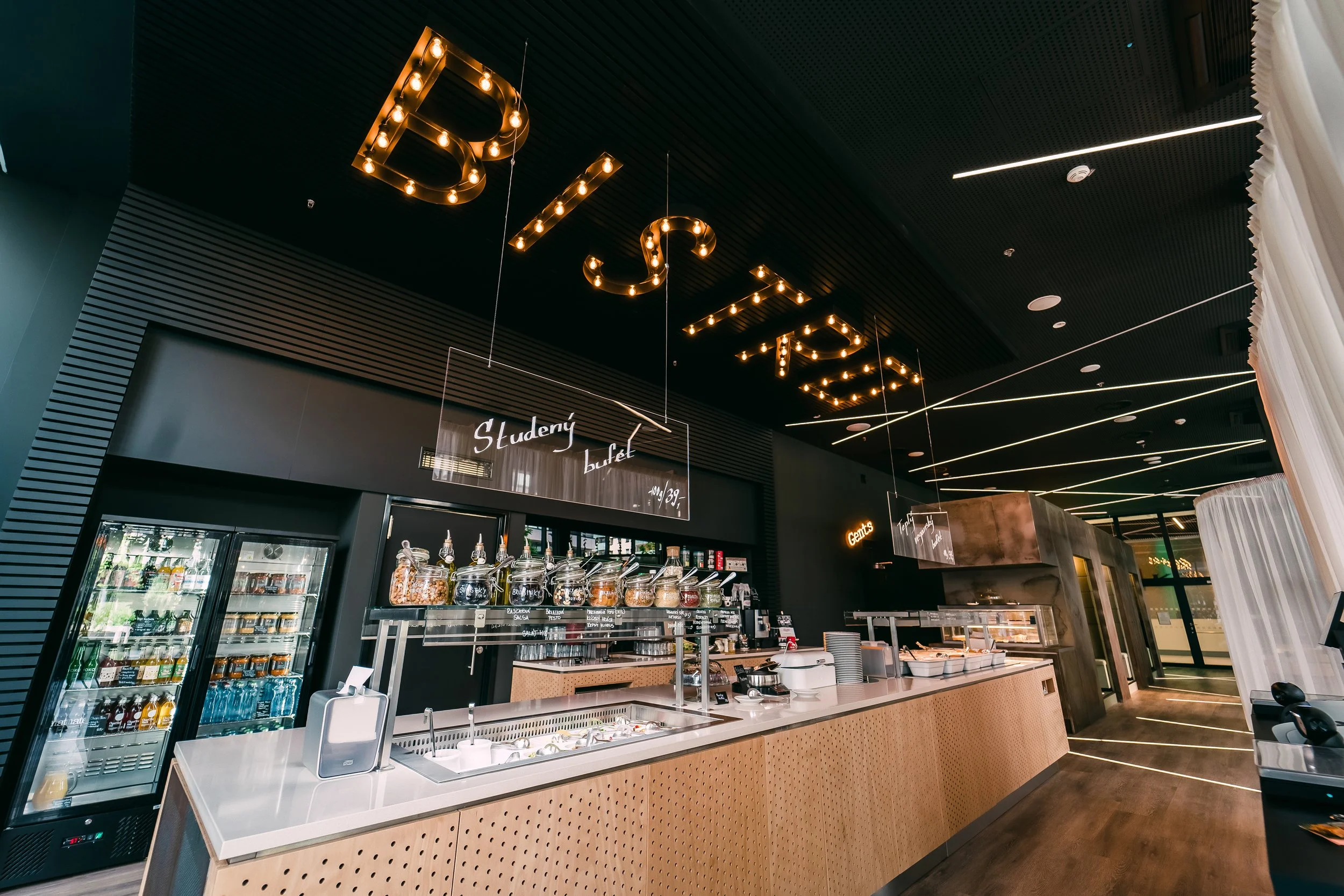 Modern cafe with black walls, illuminated spicy letter sign on ceiling, food and drink counter, refrigerators with beverages, and a decorative ceiling with linear lights. Restaurant interior photography in Zürich, Zug, Bern, Lucerne, Basel, St. Galle