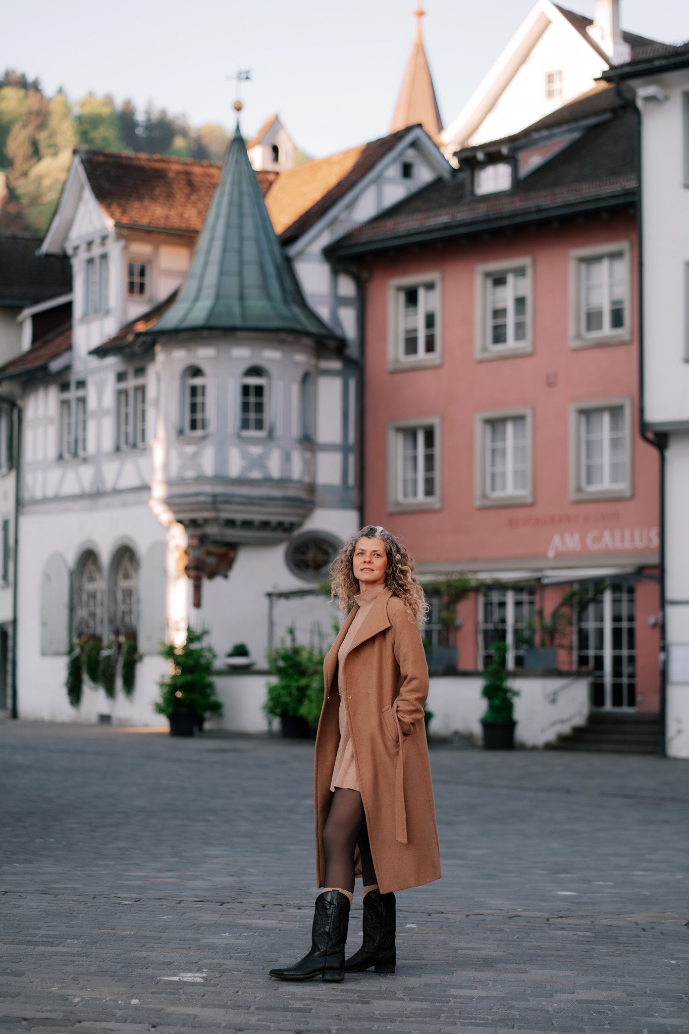 Woman in a tan coat and black cowboy boots standing on cobblestone street with colorful historic buildings of St. Gallen Switzerland in the background.
