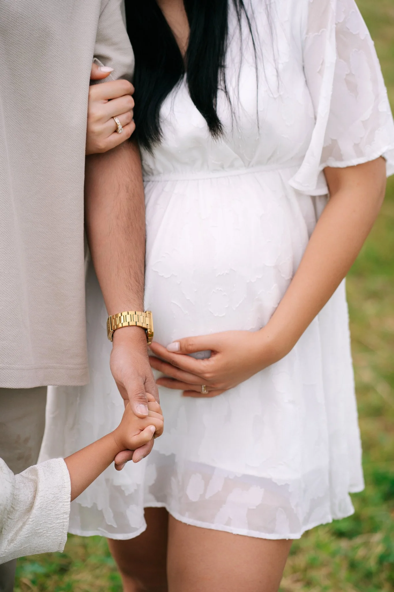 A pregnant woman wearing a white dress holds her belly while standing outdoors with a man and a child. The man is wearing a tan shirt with a gold watch, and the child in a cream sweater holds the man's hand. Maternity photoshoot in Switzerland.