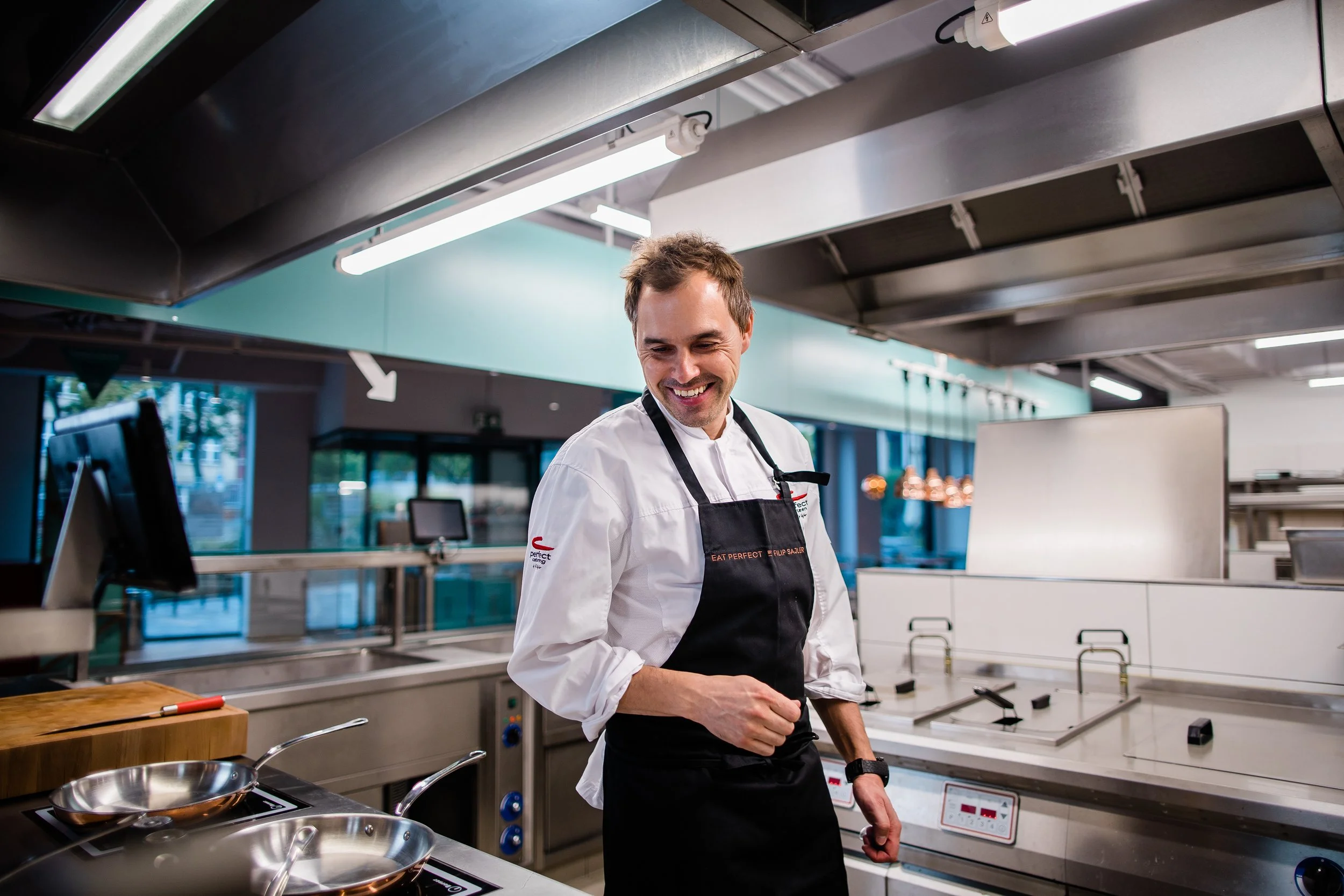 A smiling male chef in a white chef coat and black apron standing in a modern commercial kitchen with stove, pans, and stainless steel countertops. Personal branding and fine dining restaurant photography in Zürich, Switzerland.