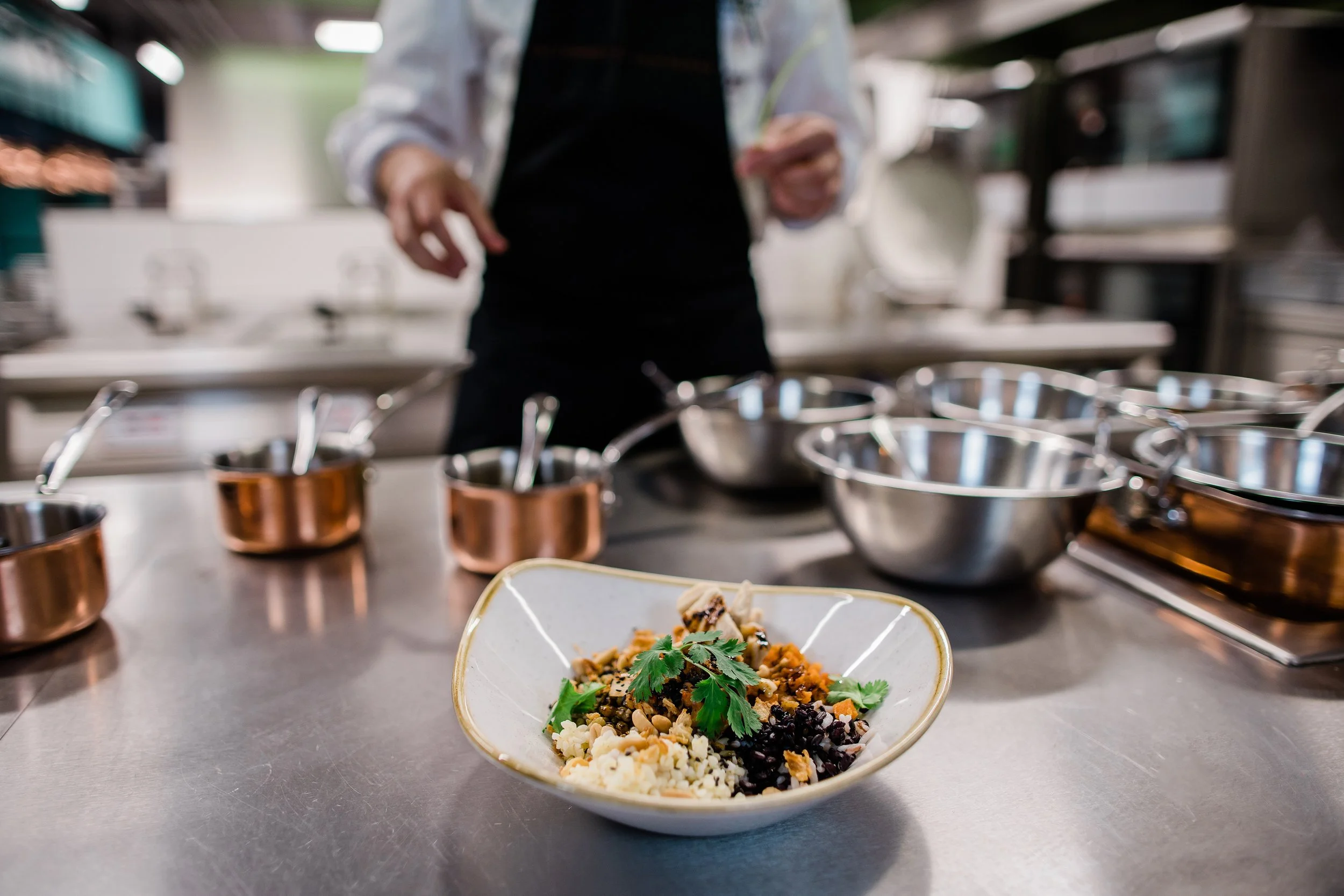 A bowl of mixed rice dish with herbs and toppings in the foreground on a kitchen counter. White apron is preparing food with various metal bowls and kitchen utensils around. Food photography fine dining luxury experience photographer Switzerland.