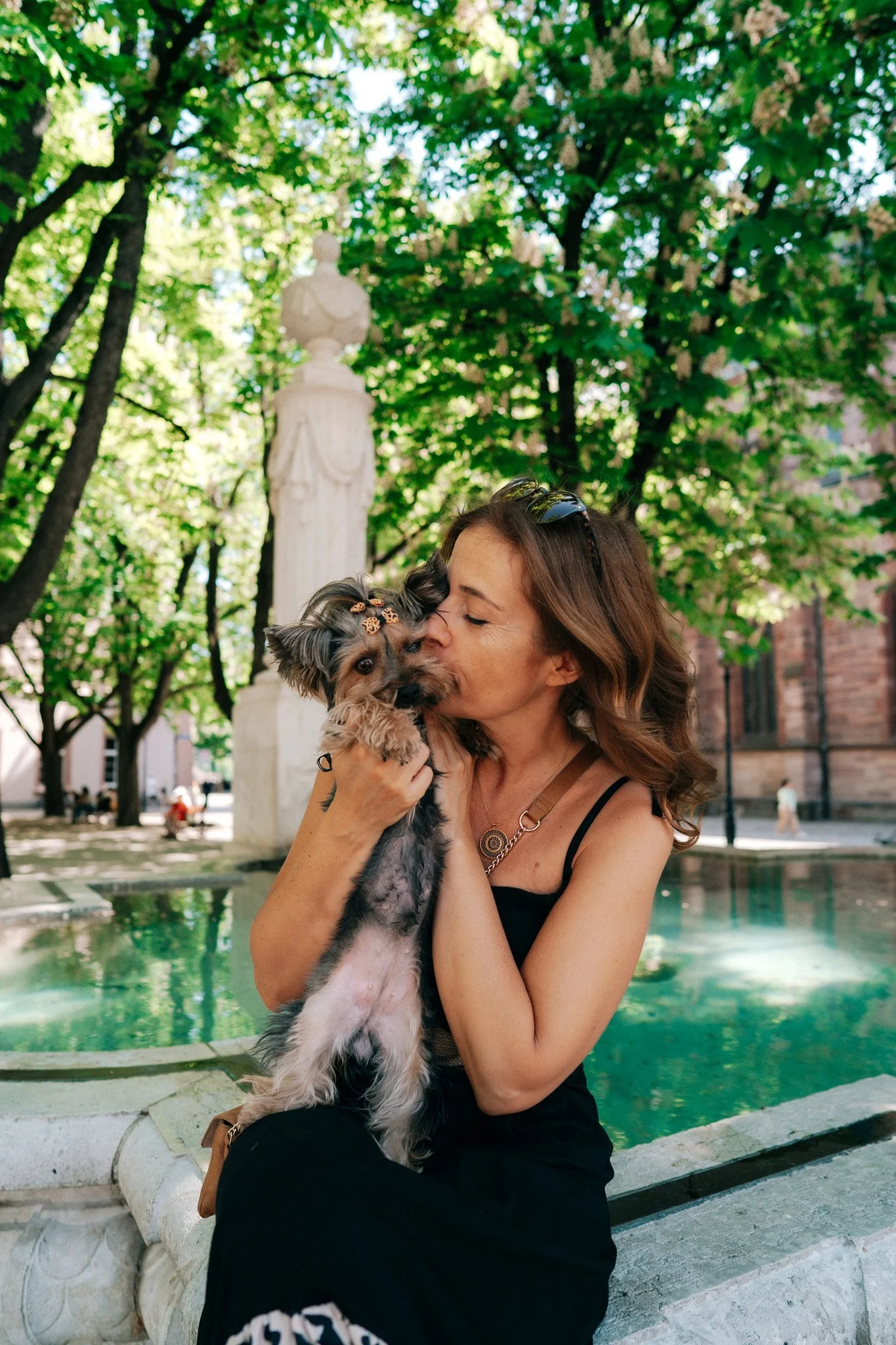 A woman with wavy brown hair, wearing a black dress with thin straps and sunglasses on her head, holds a small dog with a bow in its hair close to her face and kisses it on the cheek. They are outdoors in a park with trees, a fountain, and a historic