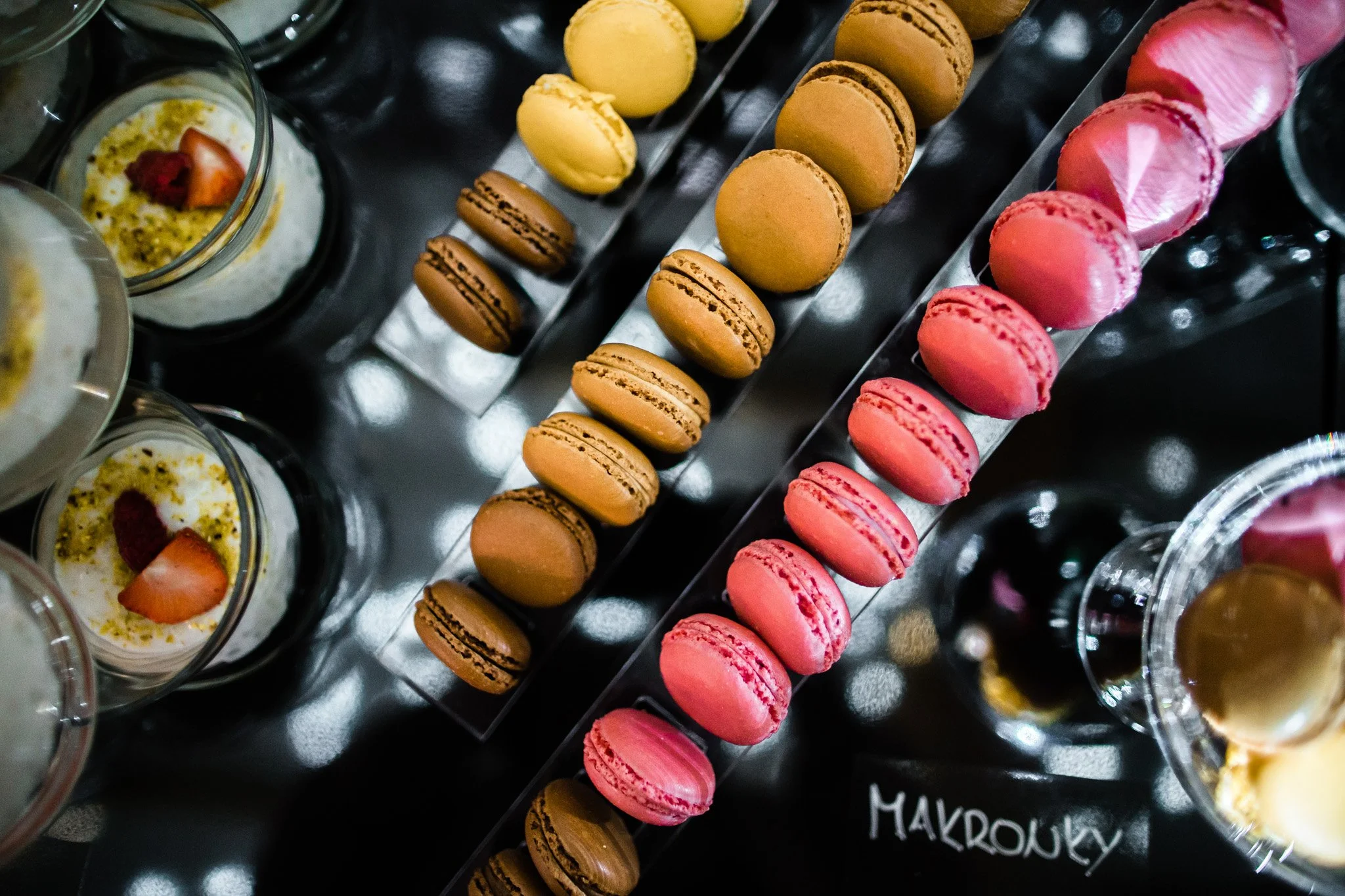 Display of colorful macarons in pink, brown, and yellow on a black surface, with some small dessert cups featuring strawberries. Catering, culinary and food photography in Zürich, Zug, Bern, Lucerne, Basel, St. Gallen and across Switzerland.