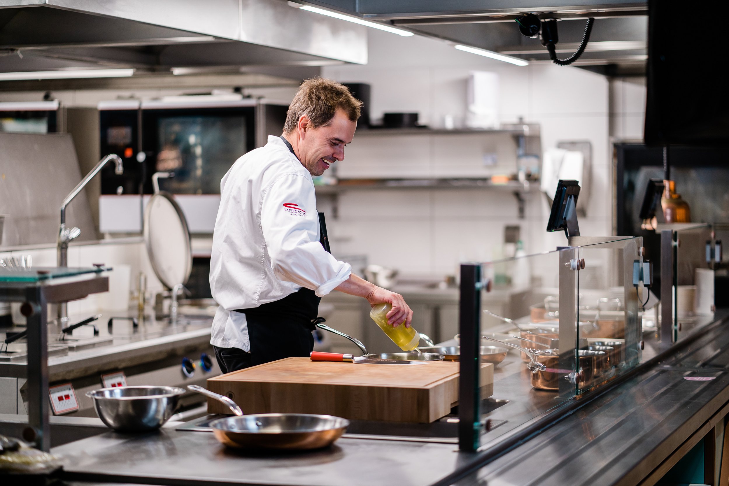 Chef adding oil in a commercial kitchen with stainless steel countertops and cooking equipment. Personal branding and fine dining restaurant photography in Zürich, Zug, Bern, Lucerne, Basel, St. Gallen and across Switzerland.