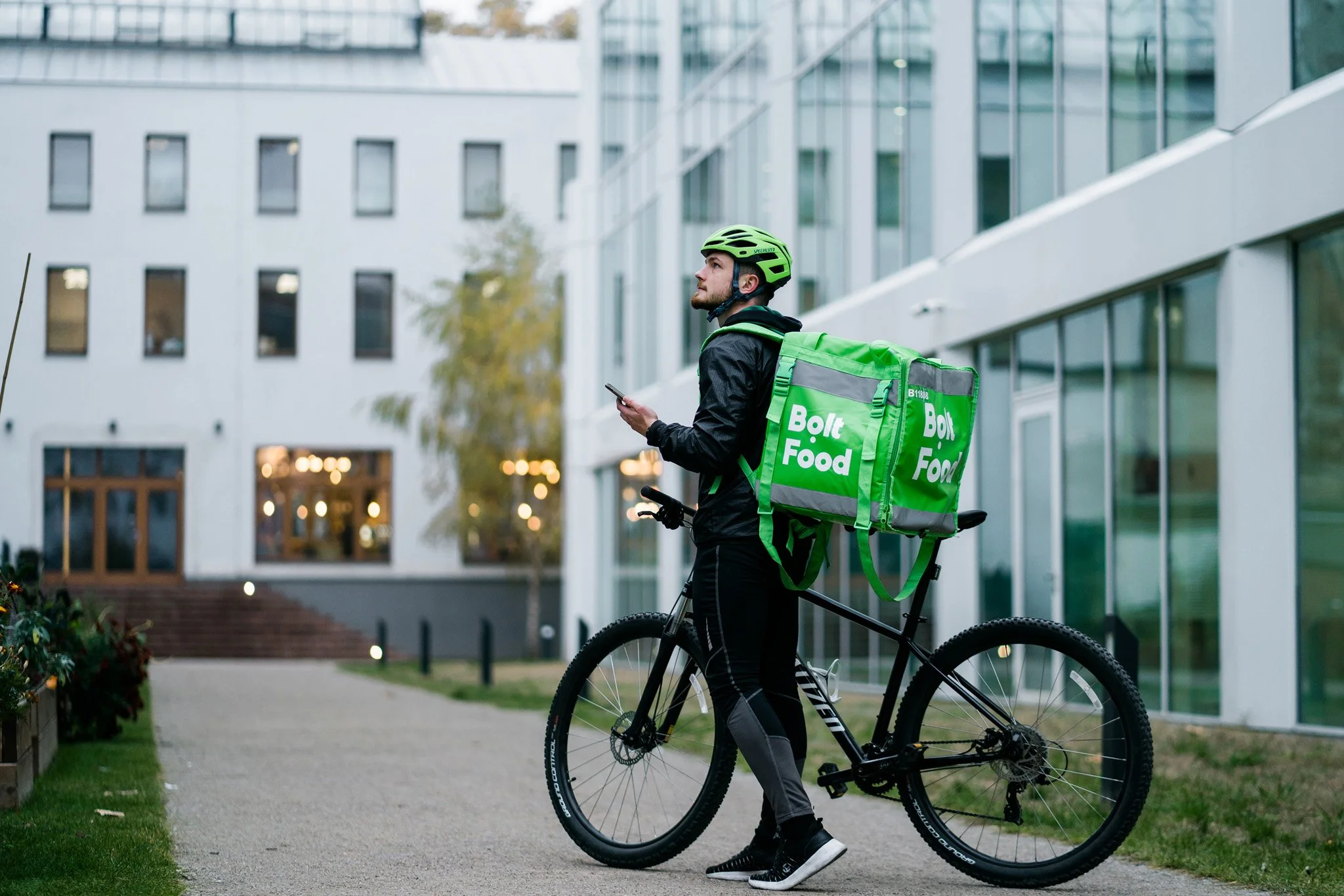 A bike delivery rider with a green helmet and a large green backpack marked 'Bolt Food' stands on a sidewalk next to his bicycle, looking at his phone, in an urban area with modern buildings. Advertising campaign photographer Switzerland.