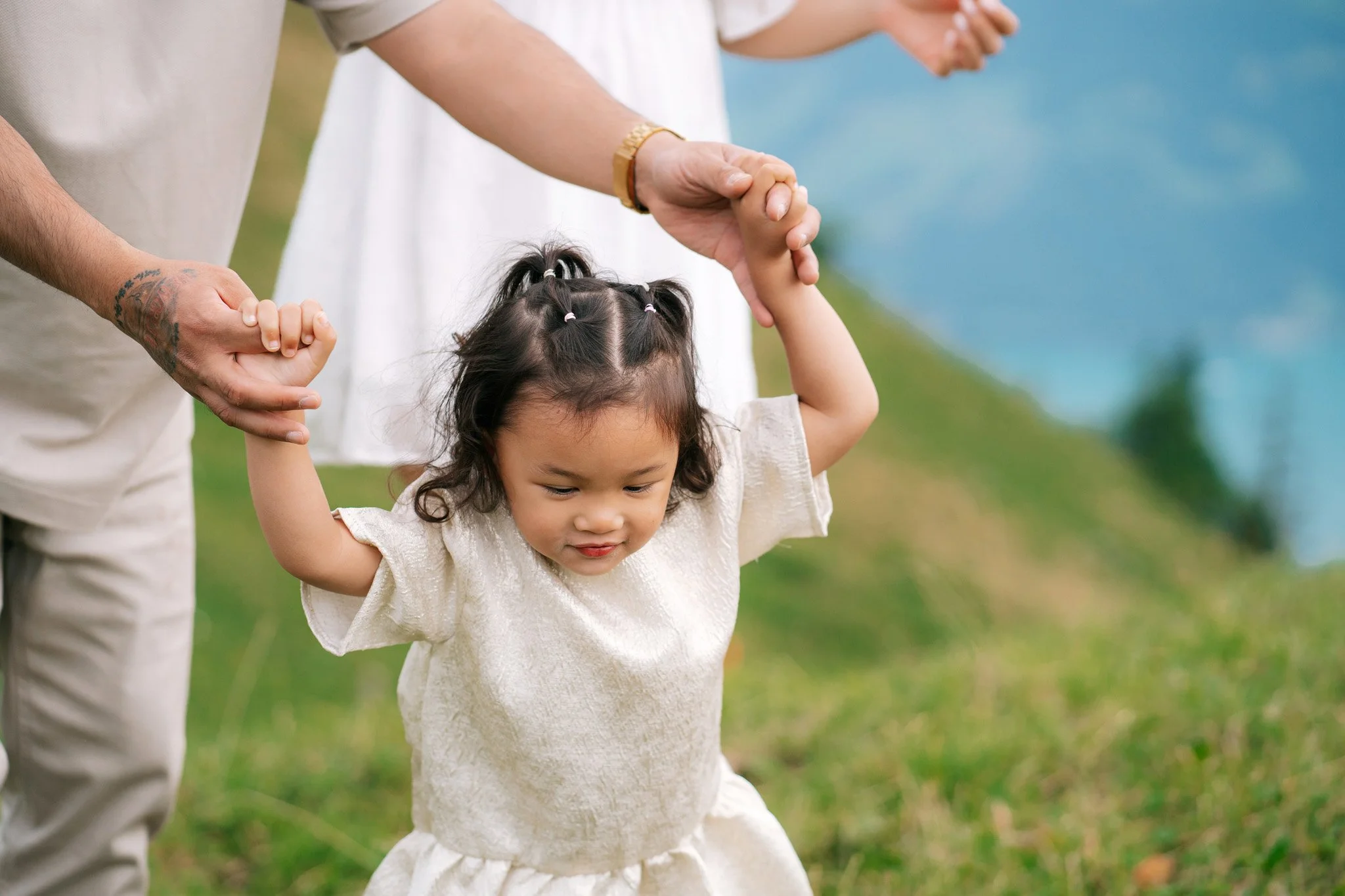 A young girl in a cream-colored dress being guided by two adults on either side as she walks outside on grassy terrain. Family photoshoot in Switzerland.