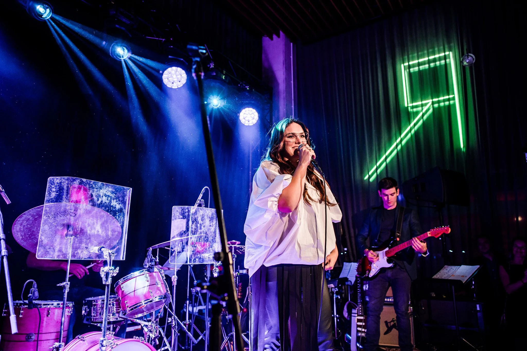 A woman singing into a microphone on stage with band members, drums, and guitar, under colorful stage lights and a neon green upward arrow sign. Concert photography in Zürich, Zug, Bern, Lucerne, Basel, St. Gallen and across Switzerland.