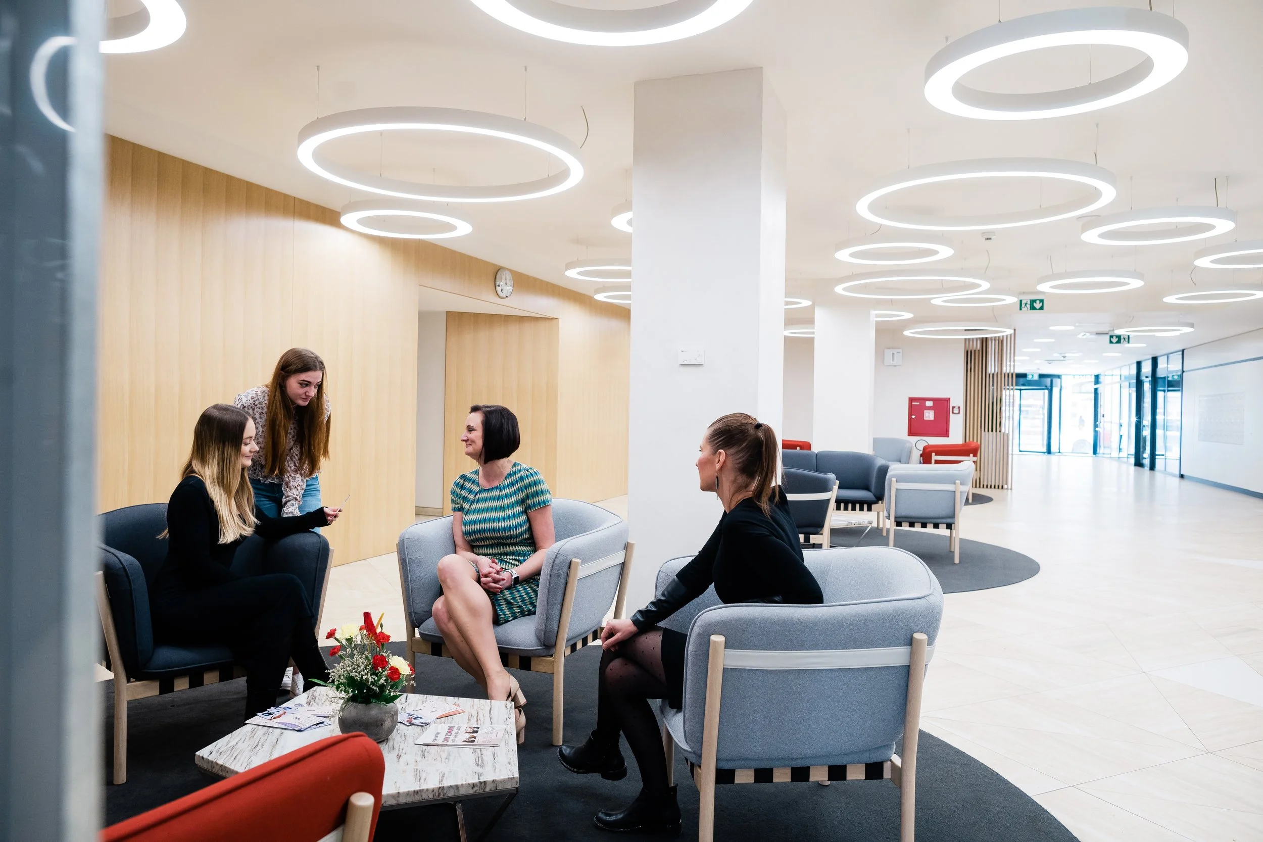 Four women sitting and standing in a modern lobby with seating, a coffee table with flowers, and circular ceiling lights. Modern light and airy corporate company employer branding photography in Switzerland - Zurich, Bern, Luzern.