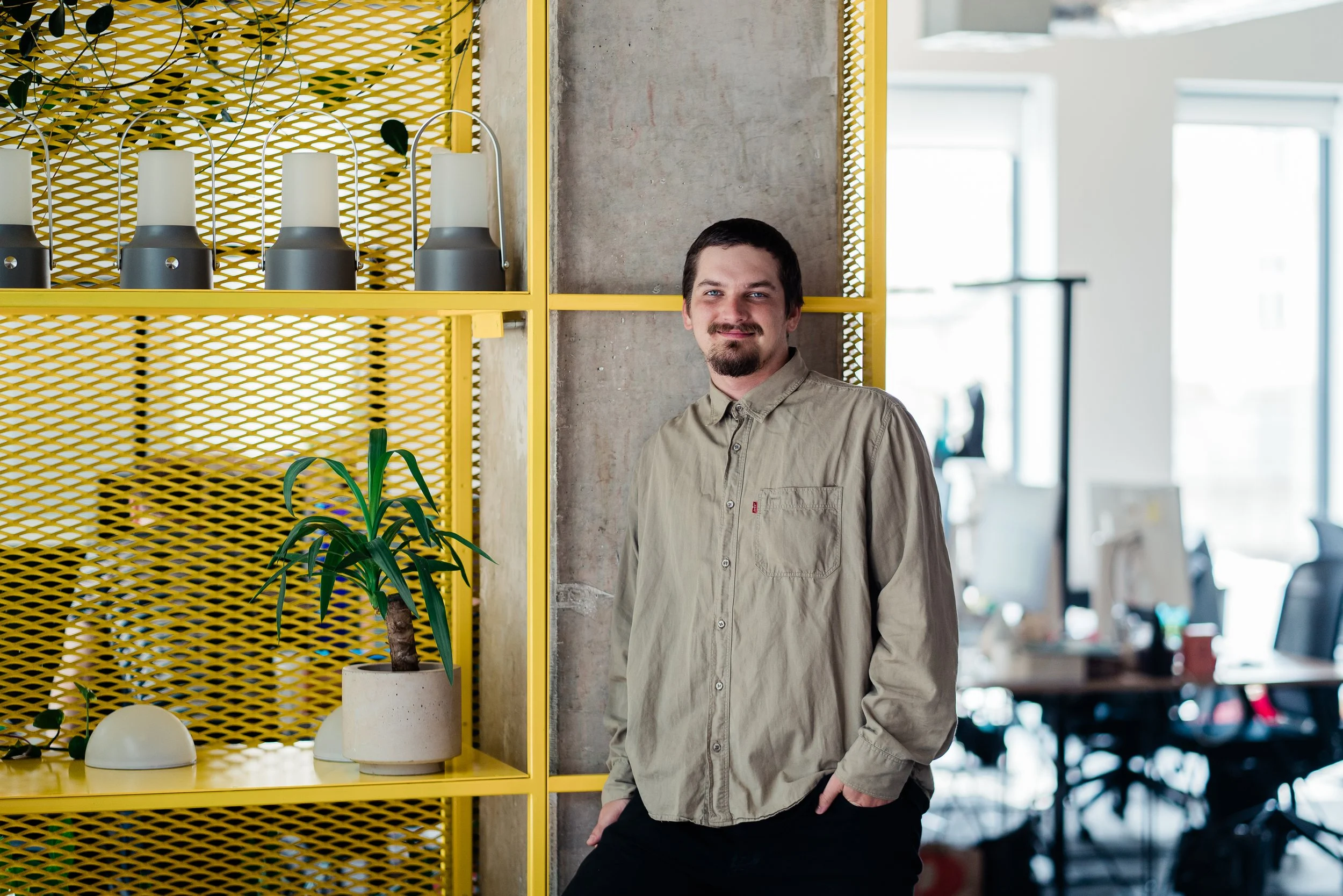 A man with dark hair and a beard wearing a beige button-up shirt, standing next to a yellow shelf with potted plants and decorative lamps, in an office with desks and computers in the background. Professional business portrait headshot Switzerland.